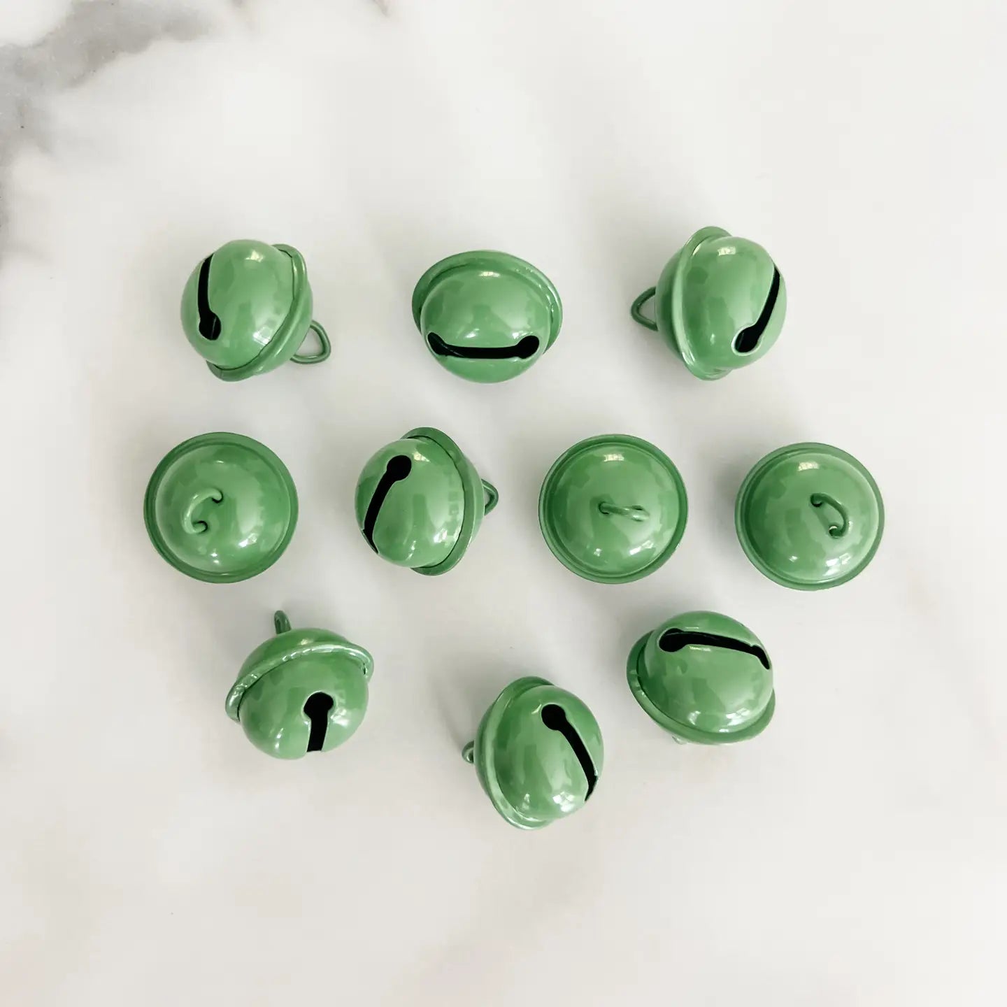Ten small glossy green jingle bells arranged in a loose circle on a light white marble surface.