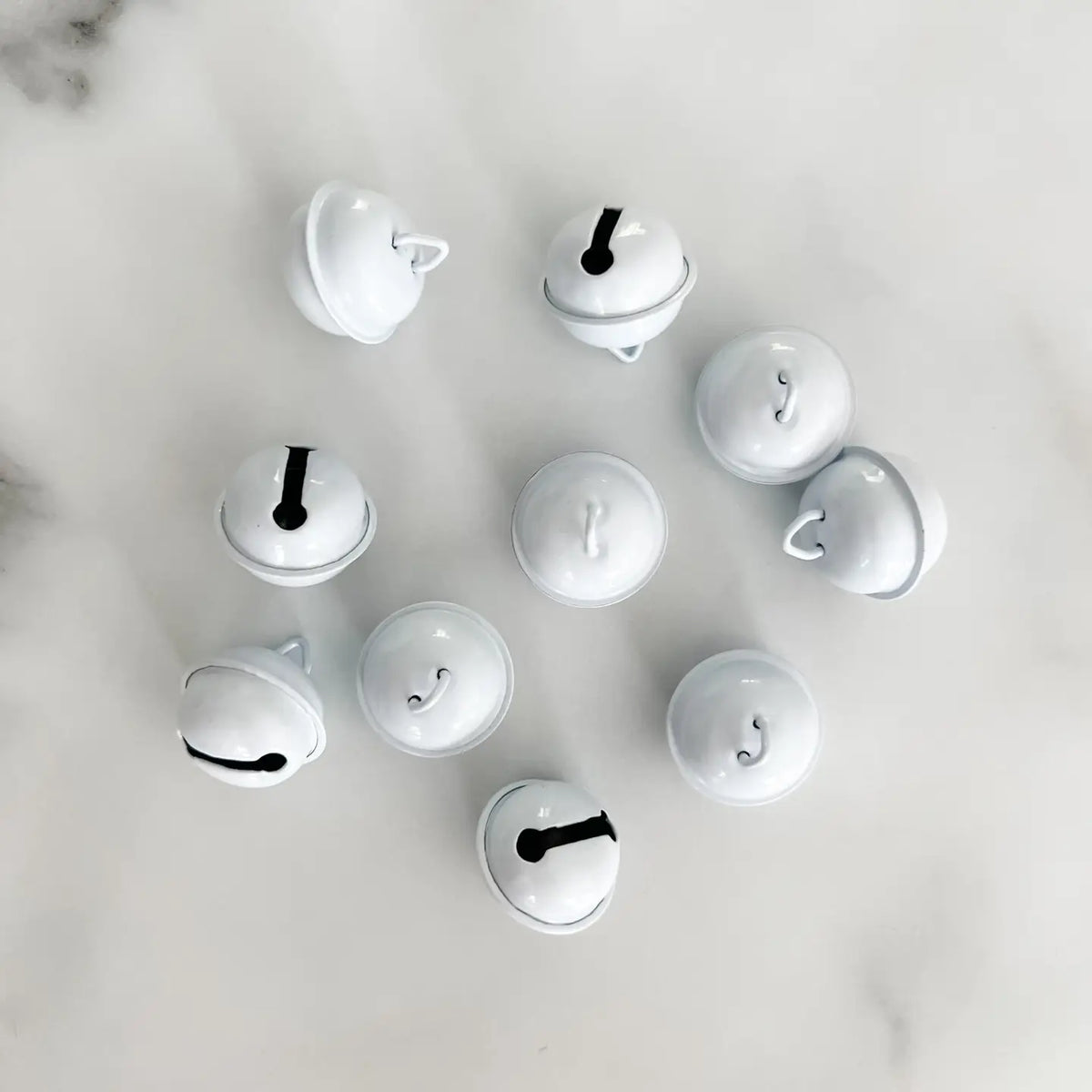 Ten small glossy white jingle bells arranged in a loose circle on a light white marble surface.
