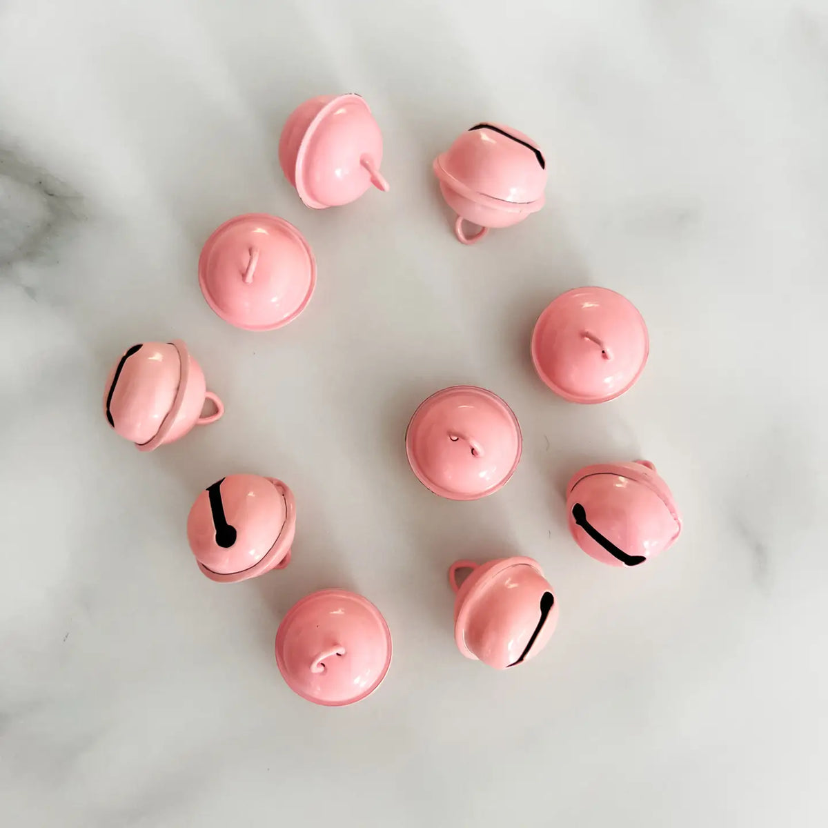 Ten small glossy pink jingle bells arranged in a loose circle on a light white marble surface.