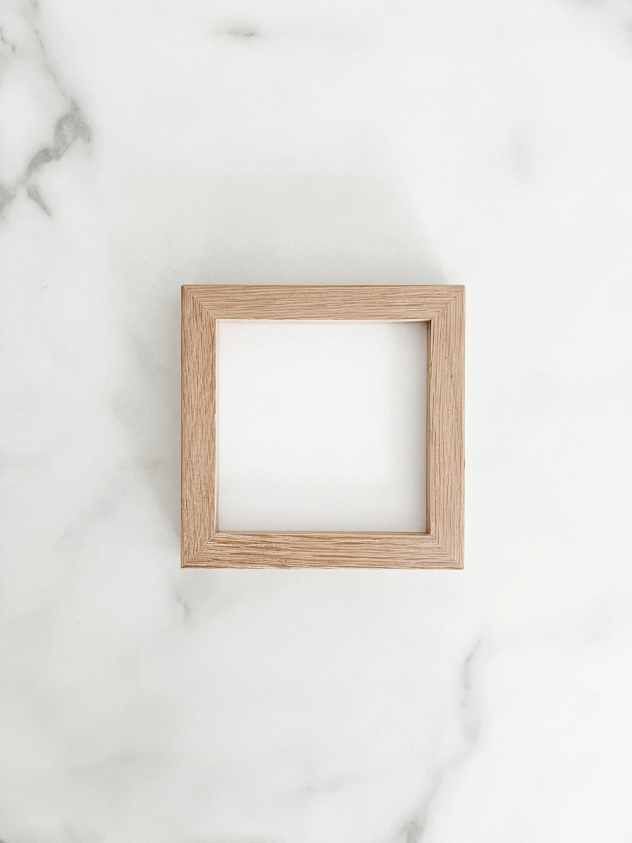 A small 4 inch by 4 inch square natural oak picture frame centered on a light marble background. 