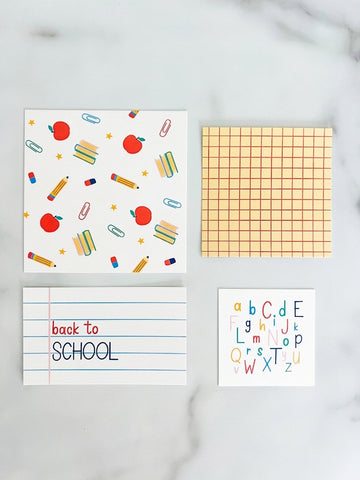 Four square back-to-school themed paper products arranged on a light marble surface: a white card with colorful school supplies and red apples, a sheet of peach grid paper, a white notepad with blue lines and the words 'back to SCHOOL' in red and blue, and a white card with colorful alphabet letters. Back to school decor inserts for tiered trays, shelves, classrooms and seasonal home decorating