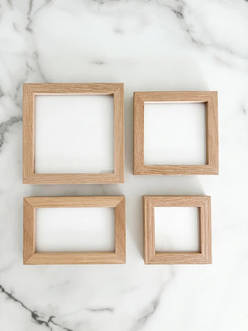 Four empty natural oak wood frames in 5x5, 4x4, 3x3, and 5x3 inch sizes arranged on a white marble background.