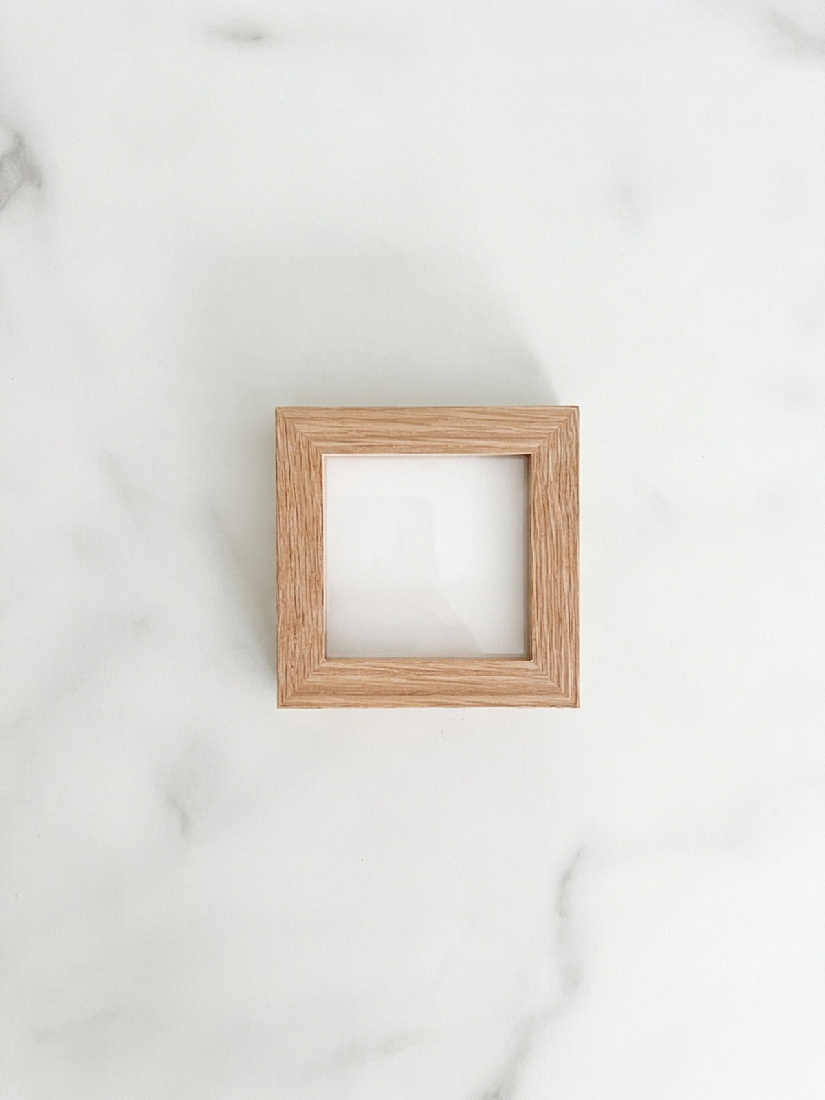 A small 3 inch by 3 inch square natural oak picture frame centered on a light marble background. 