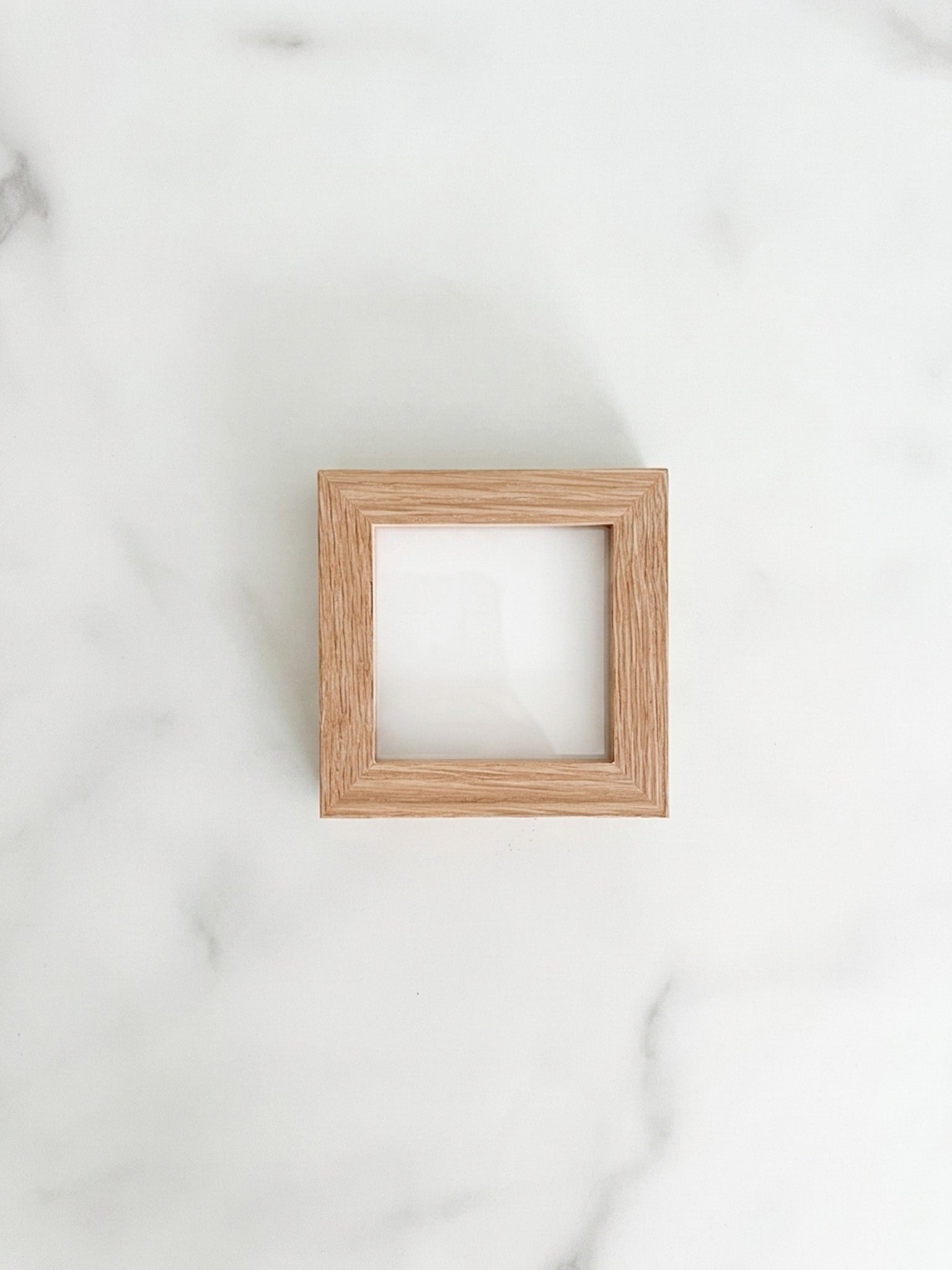 A small 3 inch by 3 inch square natural oak picture frame centered on a light marble background. 