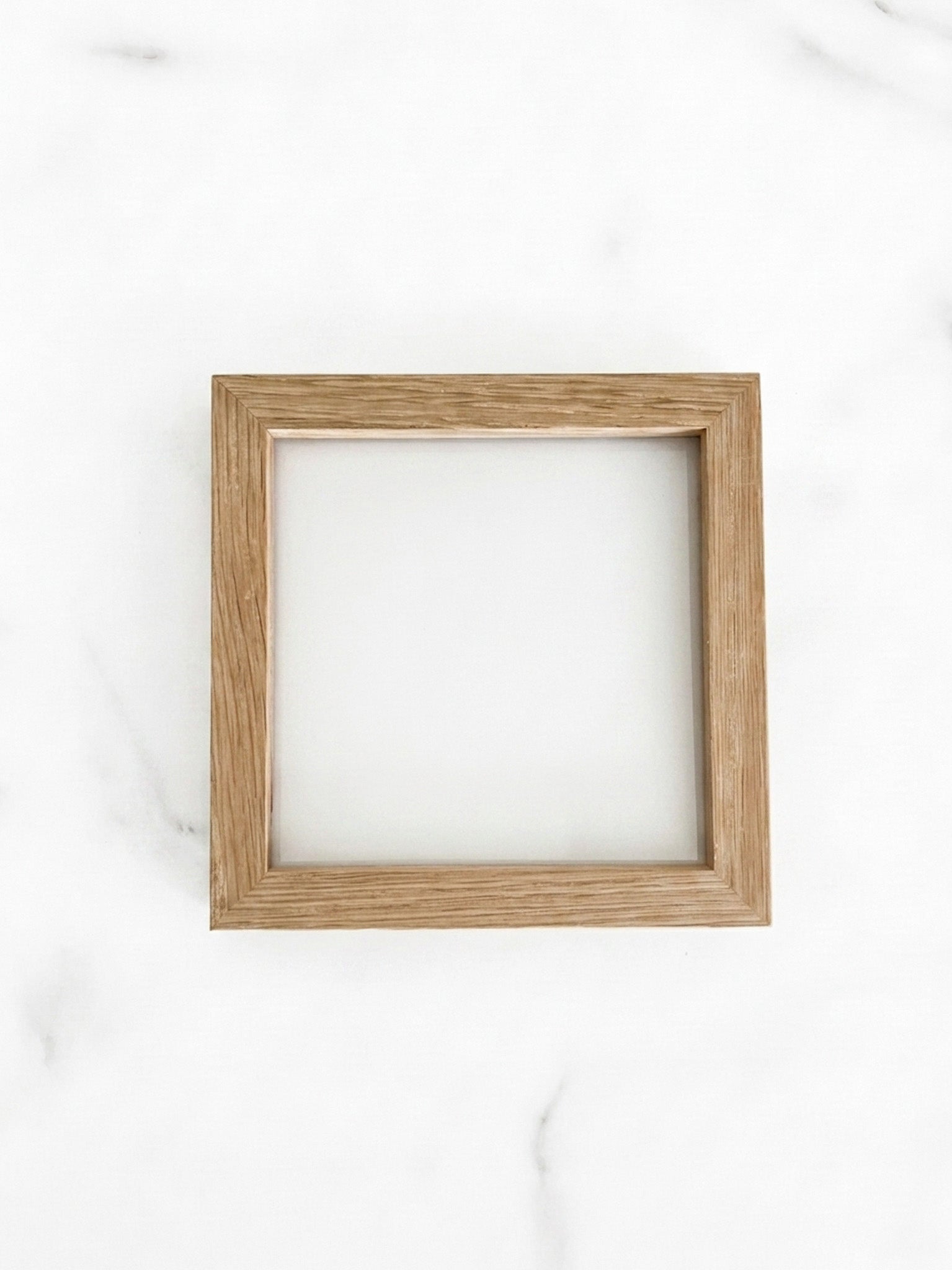 A small 5 inch by 5 inch square natural oak picture frame centered on a light marble background. 