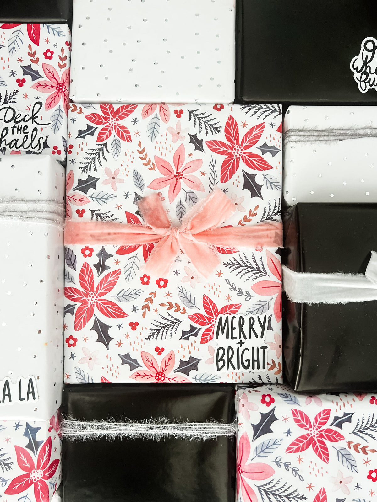 A styled flat-lay of various gift boxes wrapped in festive Christmas paper. The center box is wrapped in white paper with a red poinsettia and gray holly pattern, tied with a pink velvet ribbon.
