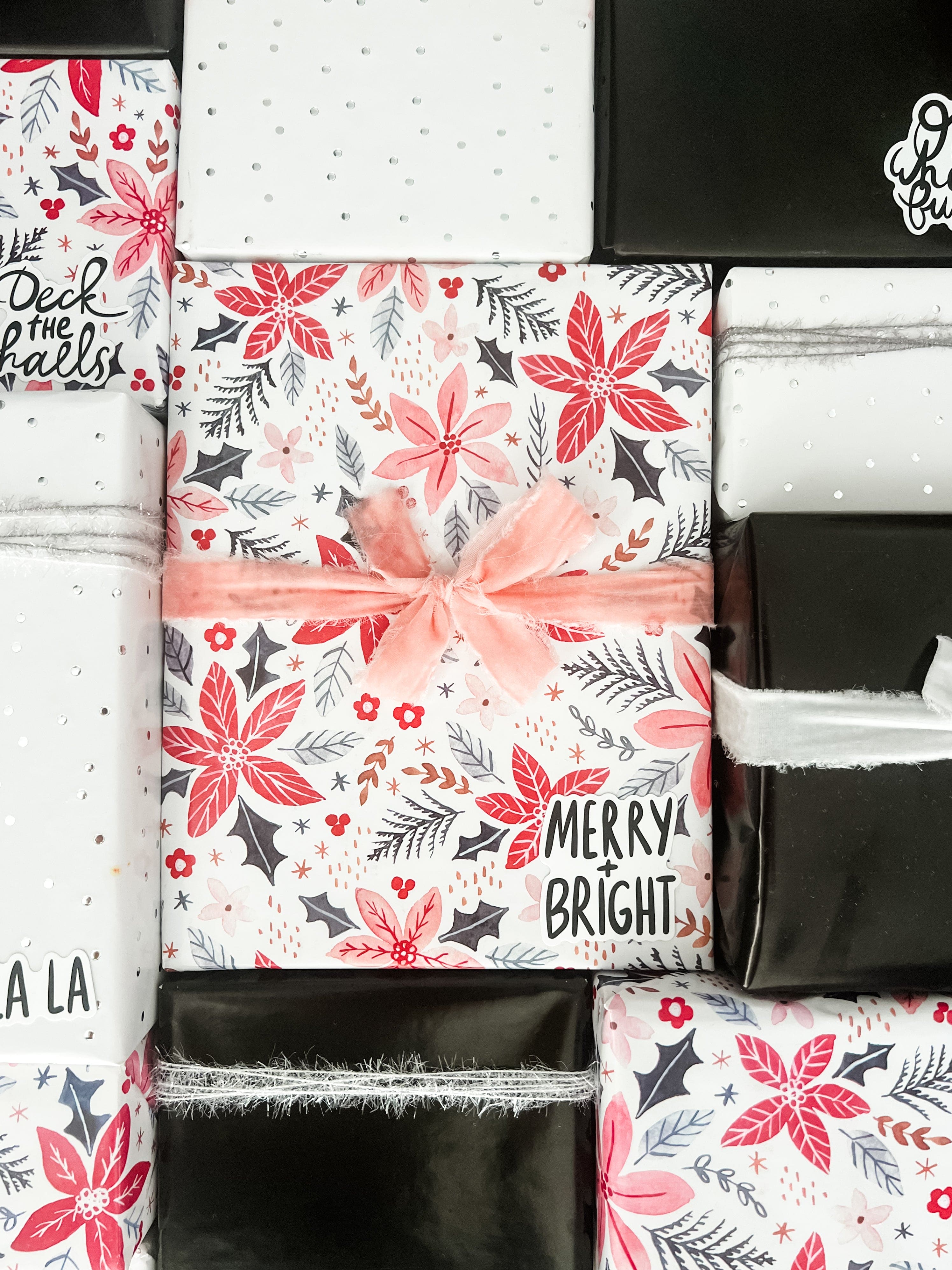 A styled flat-lay of various gift boxes wrapped in festive Christmas paper. The center box is wrapped in white paper with a red poinsettia and gray holly pattern, tied with a pink velvet ribbon.