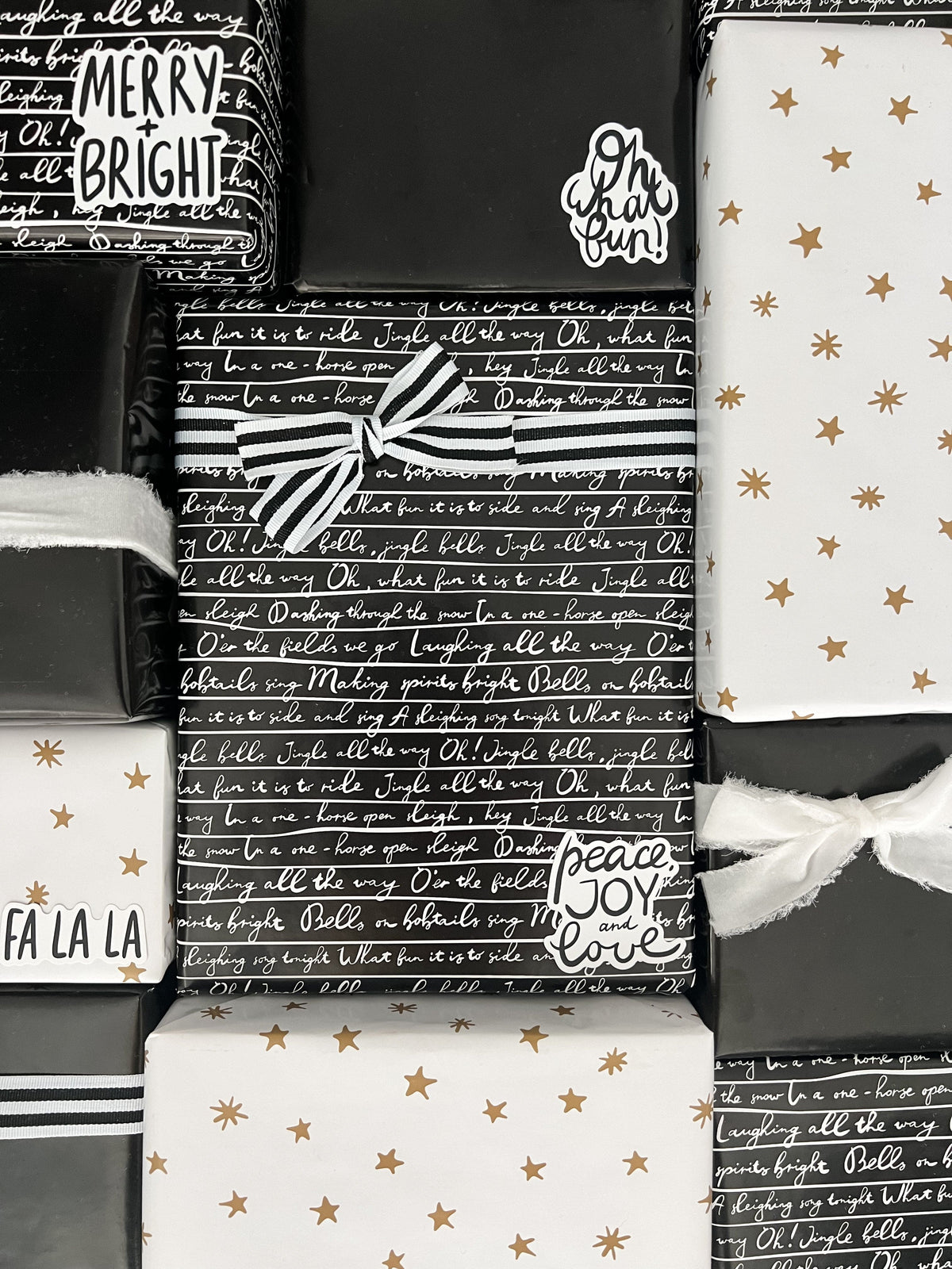 A flat-lay arrangement of wrapped gift boxes and gift bags featuring holiday-themed wrapping paper designs. The designs include black and white script patterns, white with gold stars, and black and white stripes with white ribbons.