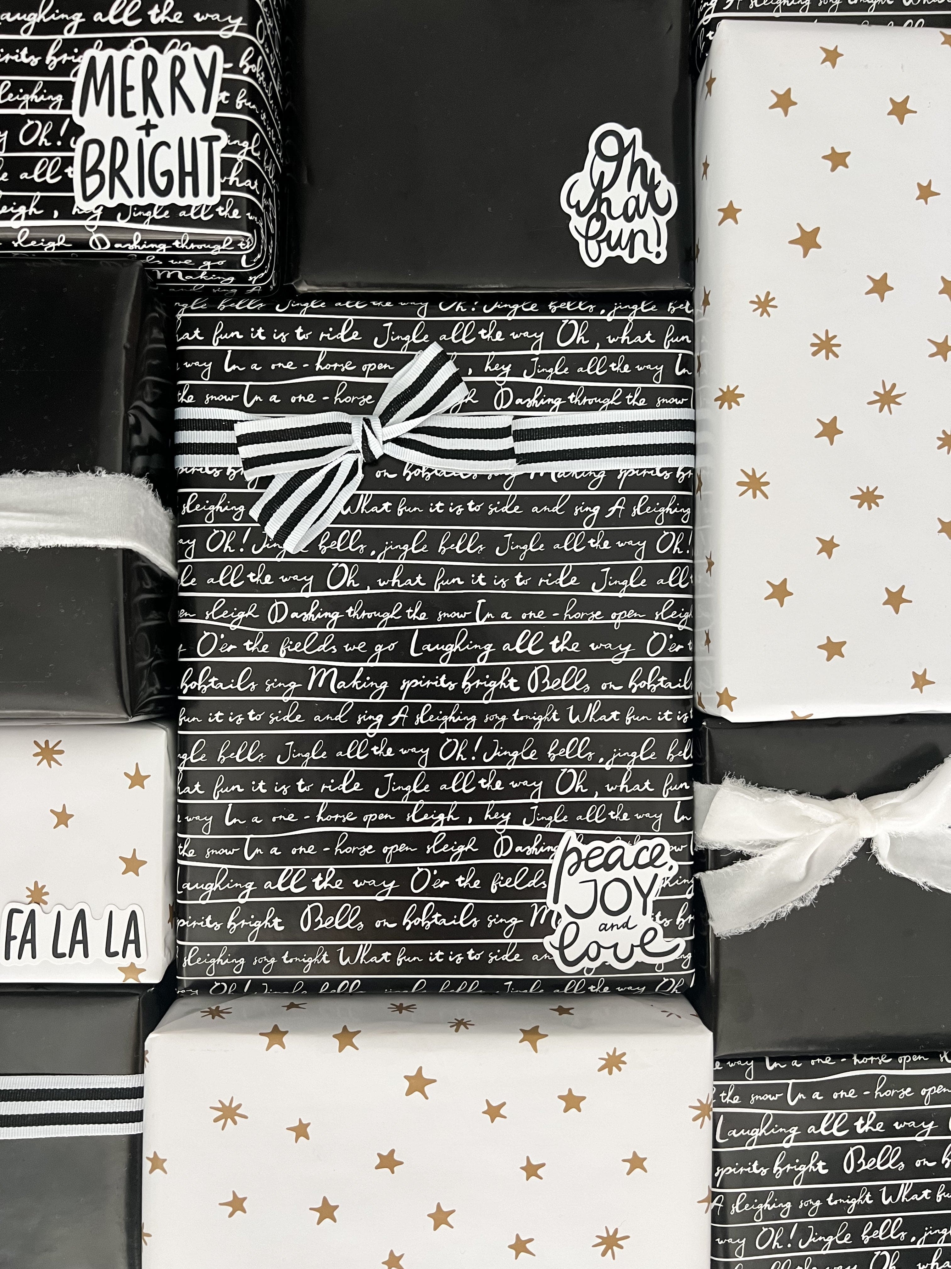 A flat-lay arrangement of wrapped gift boxes and gift bags featuring holiday-themed wrapping paper designs. The designs include black and white script patterns, white with gold stars, and black and white stripes with white ribbons.