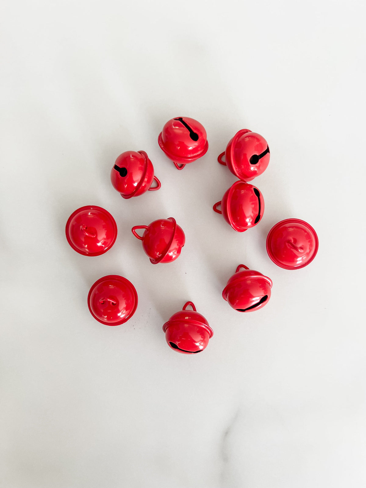 Ten small glossy red jingle bells arranged in a loose circle on a light white marble surface.