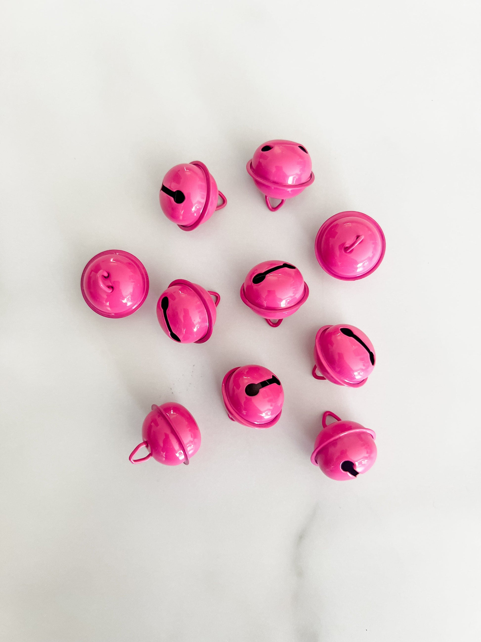 Ten small glossy raspberry, pink, jingle bells arranged in a loose circle on a light white marble surface.