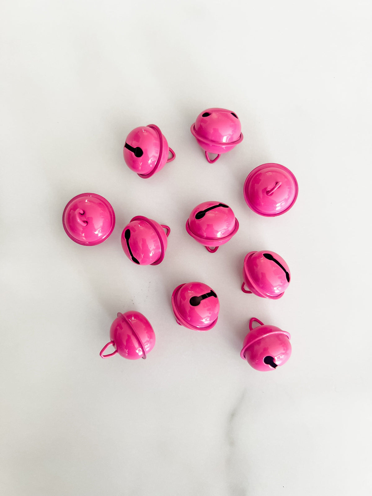 Ten small glossy raspberry, pink, jingle bells arranged in a loose circle on a light white marble surface.