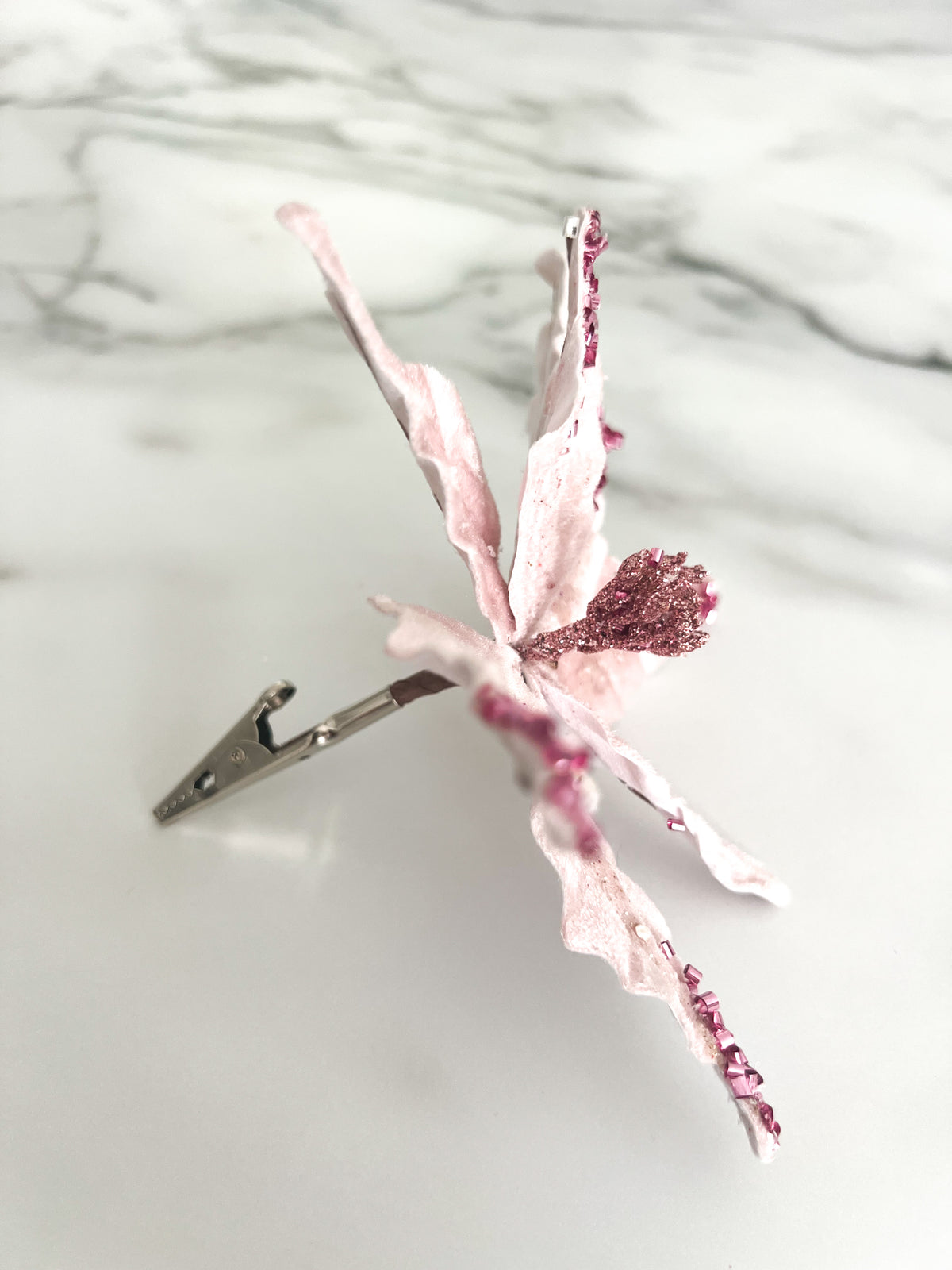 A pink beaded poinsettia flower accessory displayed on a white marble surface with a silver clip.