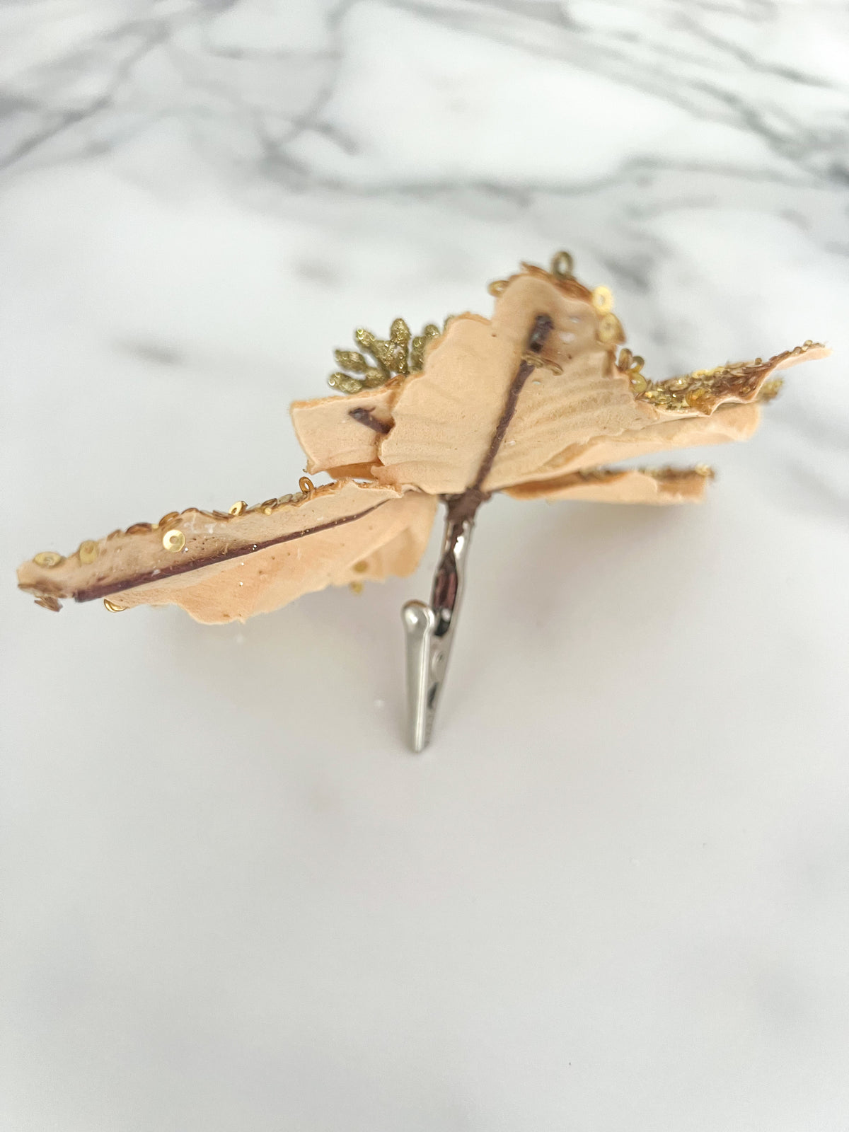 The back side of a gold sequined poinsettia flower accessory displayed on a white marble surface with a silver clip.