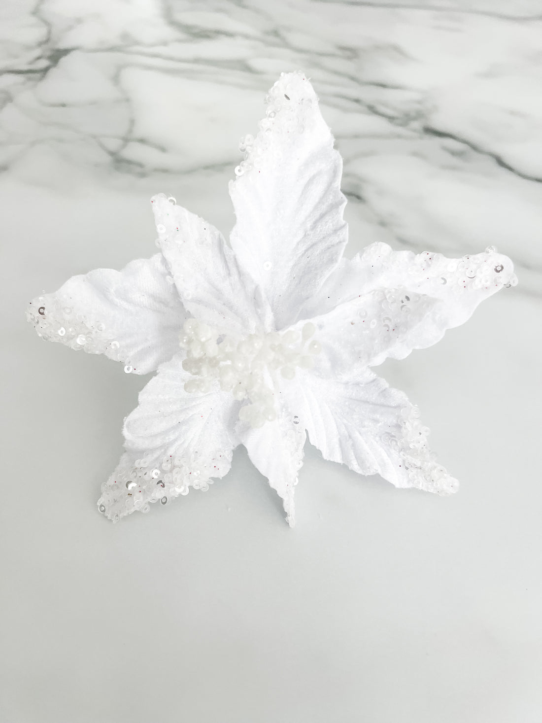 A white faux floral ornament or decorative topper with textured, glittery petals and small bead details, displayed on a light gray marble-patterned surface.