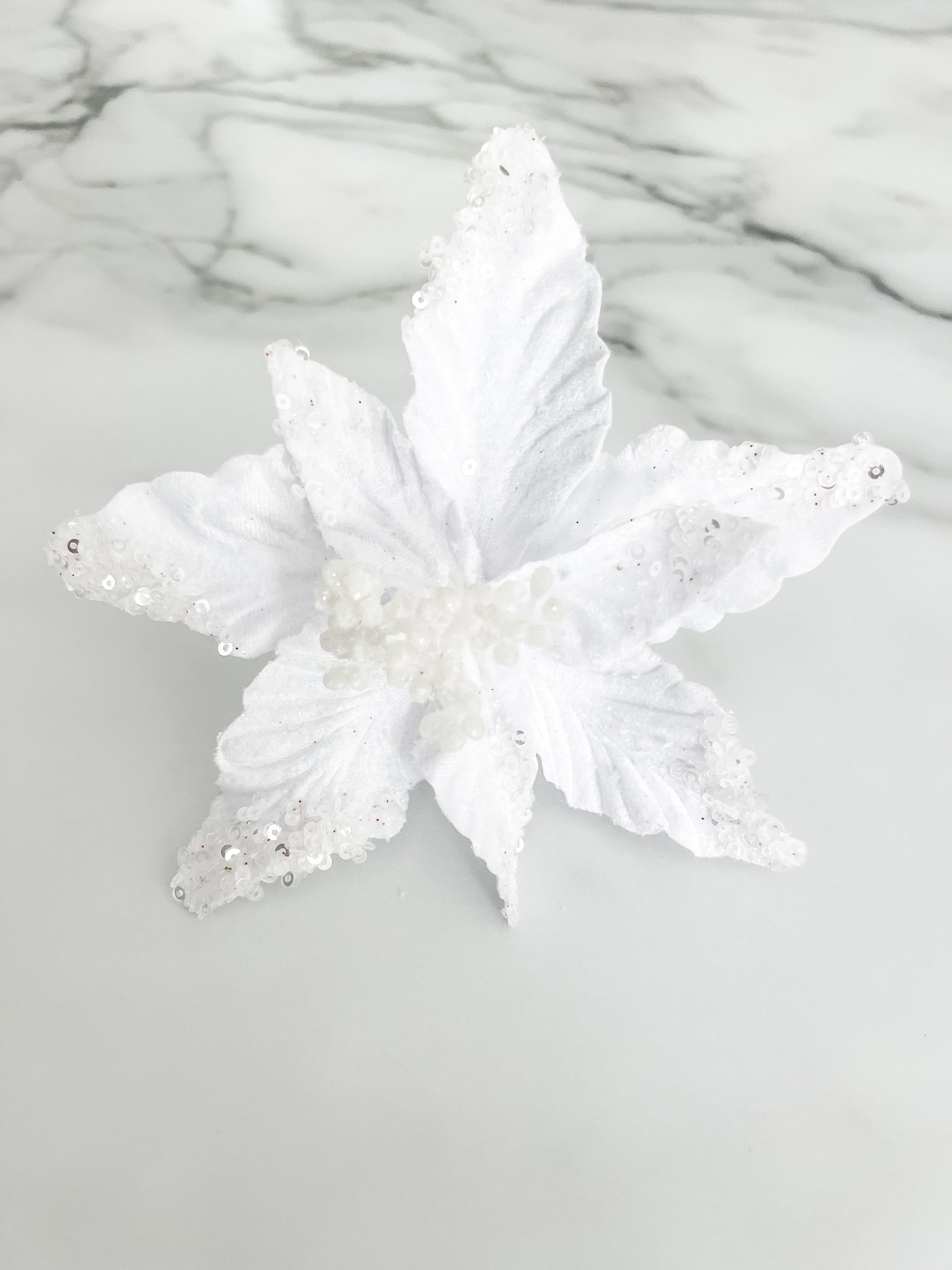 A white faux floral ornament or decorative topper with textured, glittery petals and small bead details, displayed on a light gray marble-patterned surface.