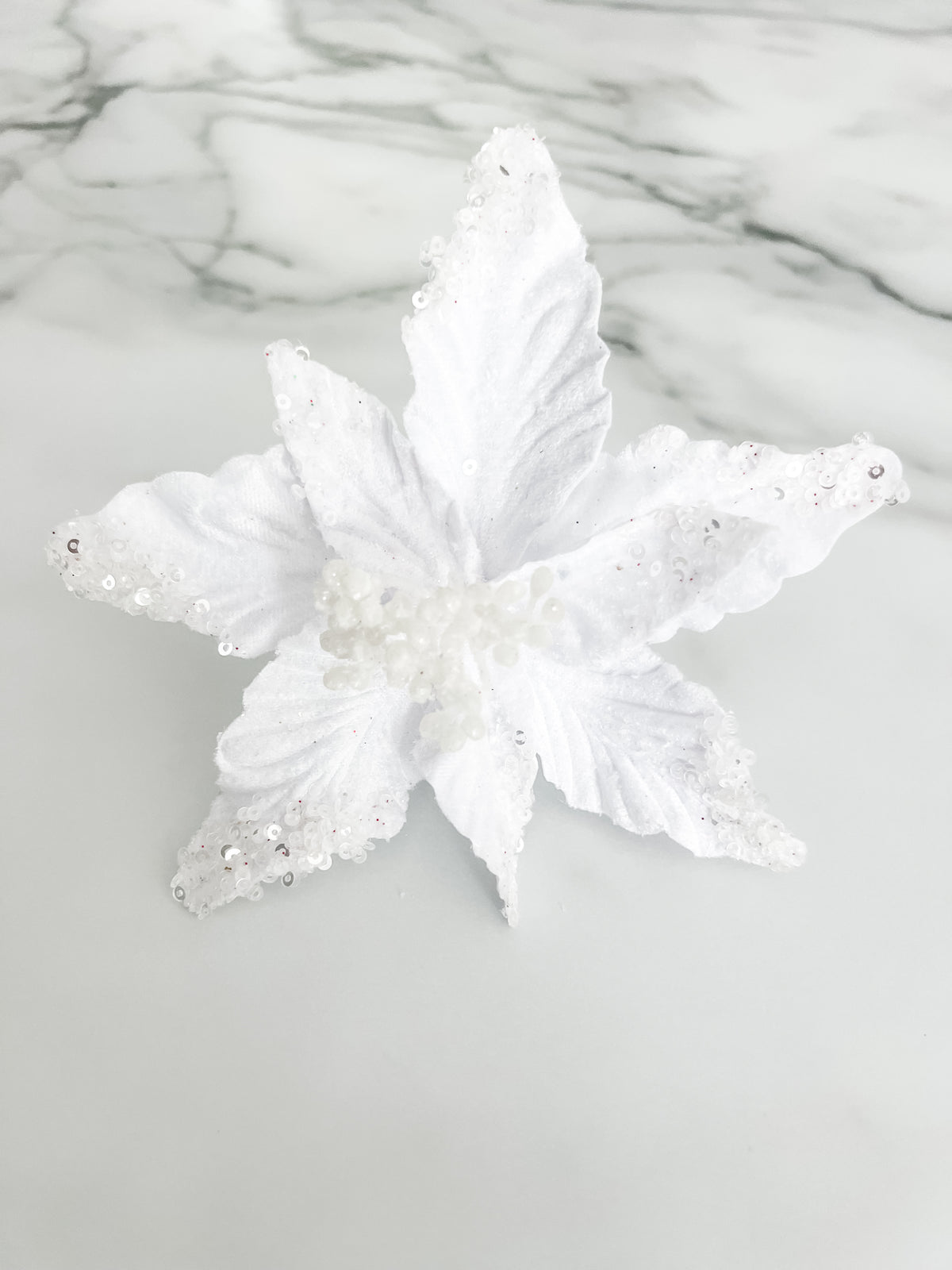 A white faux floral ornament or decorative topper with textured, glittery petals and small bead details, displayed on a light gray marble-patterned surface.