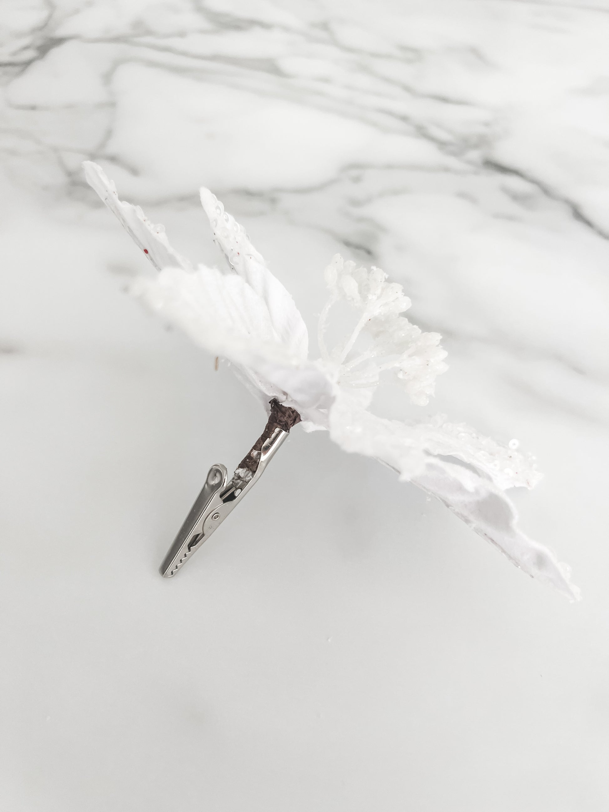A white faux floral ornament or decorative topper with textured, glittery petals and small bead details and a silver clip on the back, displayed on a light gray marble-patterned surface.