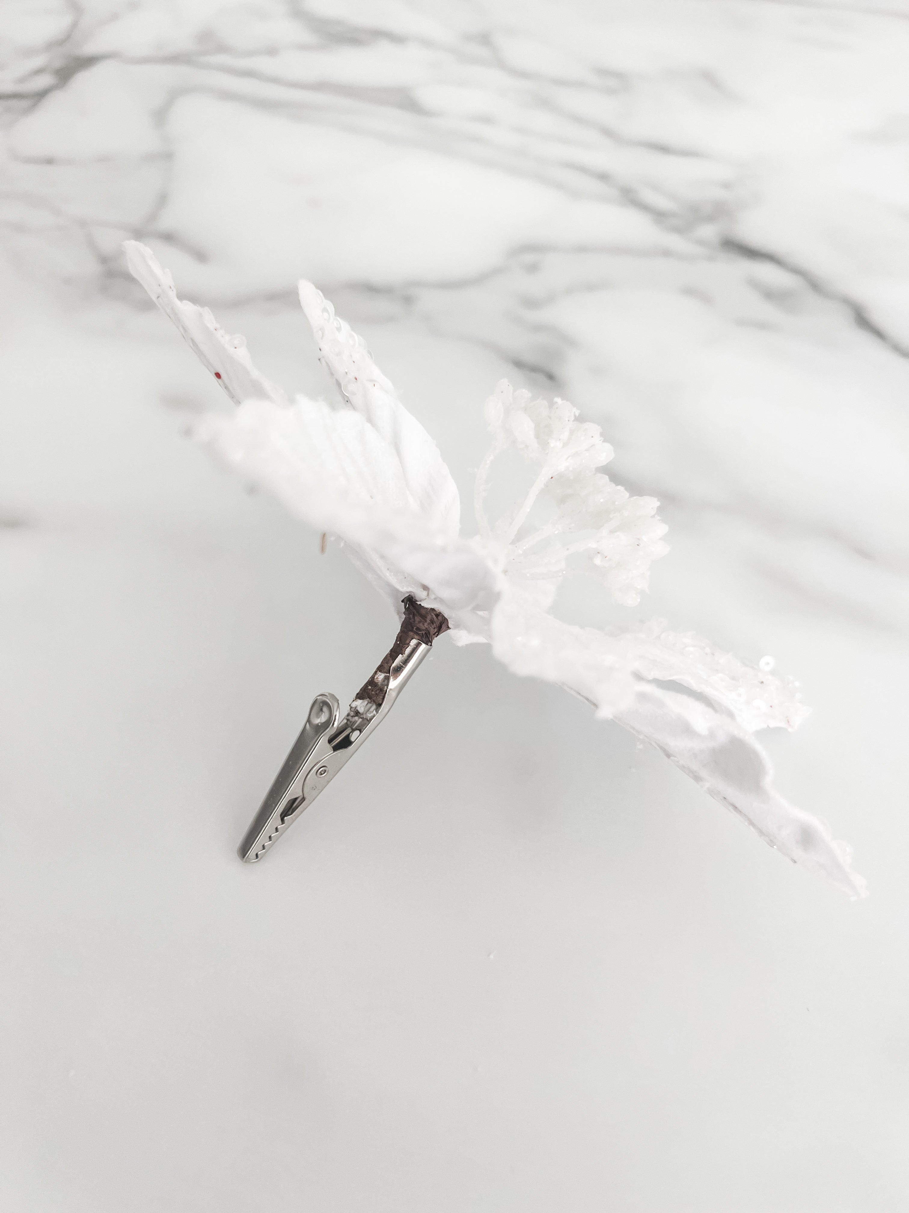 A white faux floral ornament or decorative topper with textured, glittery petals and small bead details and a silver clip on the back, displayed on a light gray marble-patterned surface.