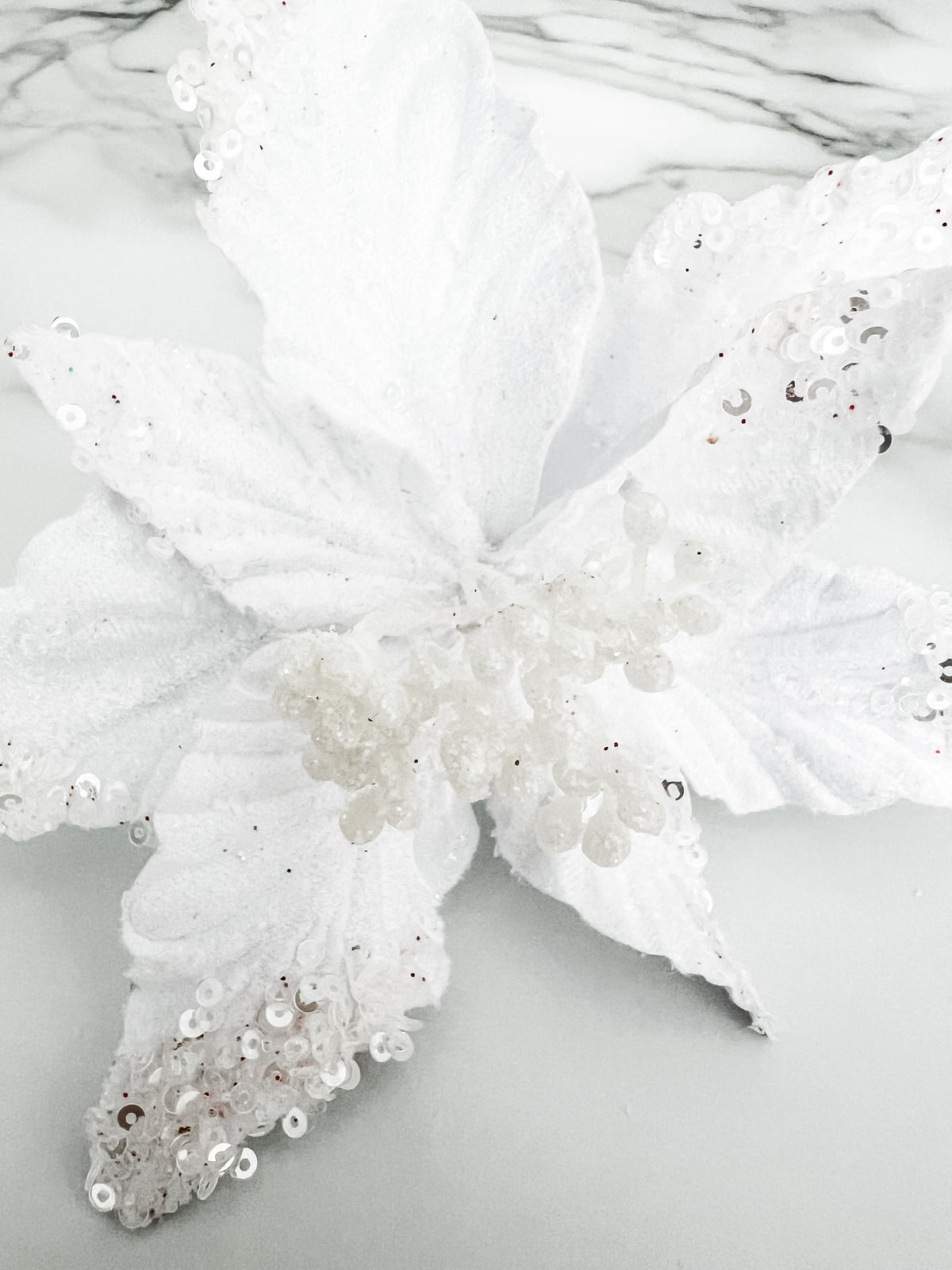 A white artificial poinsettia-style flower decoration with textured fabric petals, clear rhinestones, and silver sequins, displayed on a light marble surface.