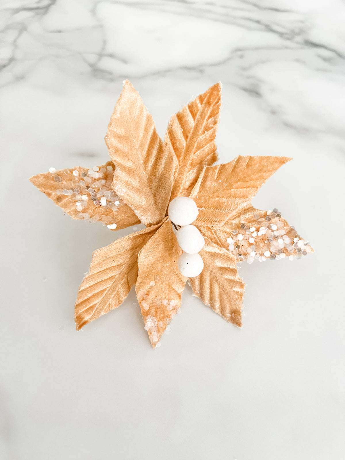 A gold velvet poinsettia-style holiday decoration with textured leaves, white faux berries, and silver glitter accents, photographed on a white marble surface.