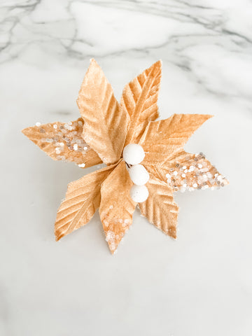 A gold velvet poinsettia-style holiday decoration with textured leaves, white faux berries, and silver glitter accents, photographed on a white marble surface.