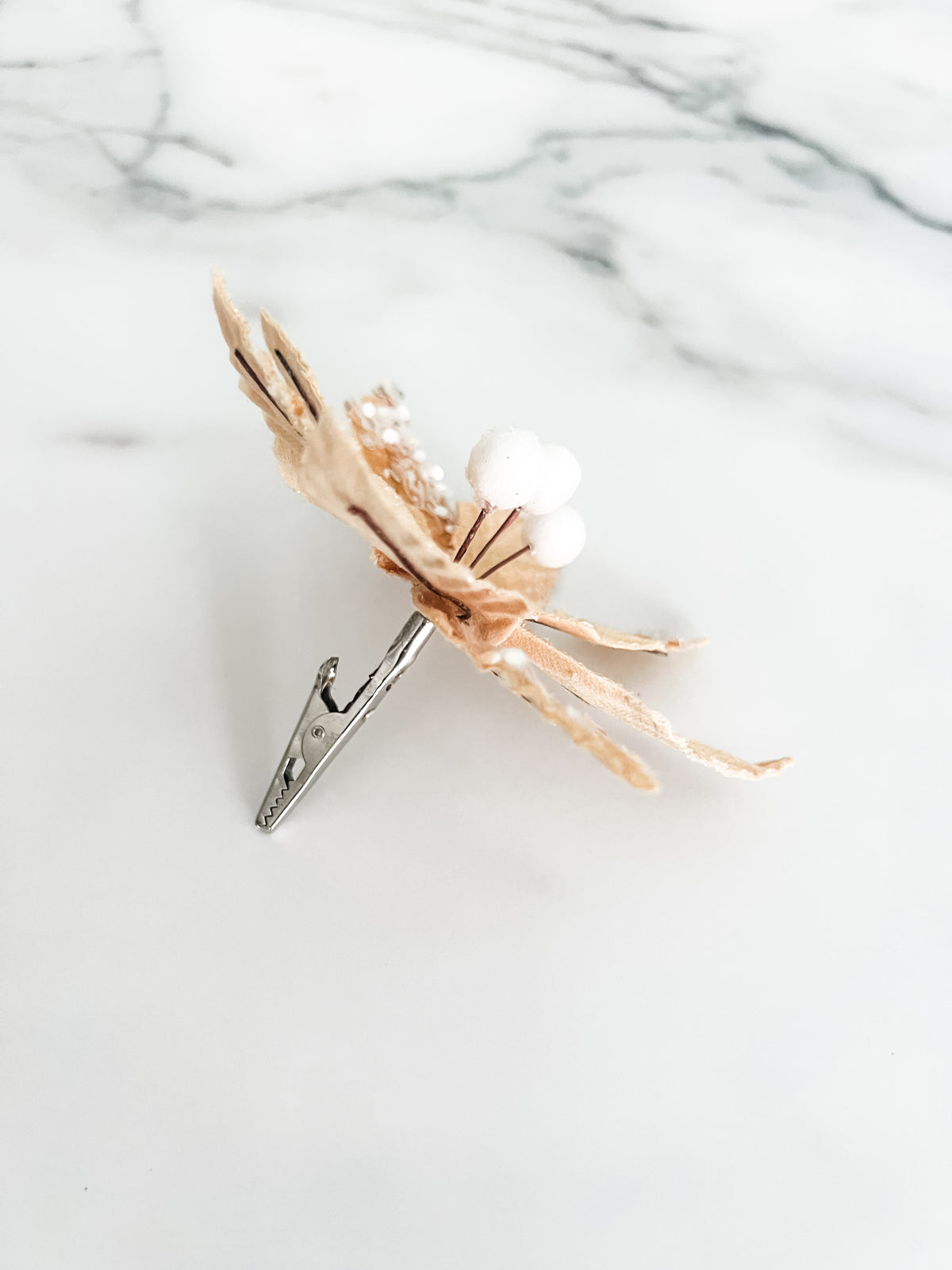 A gold velvet poinsettia-style holiday decoration with textured leaves, white faux berries, and silver glitter accents, photographed on a white marble surface with a silver clip on the back.
