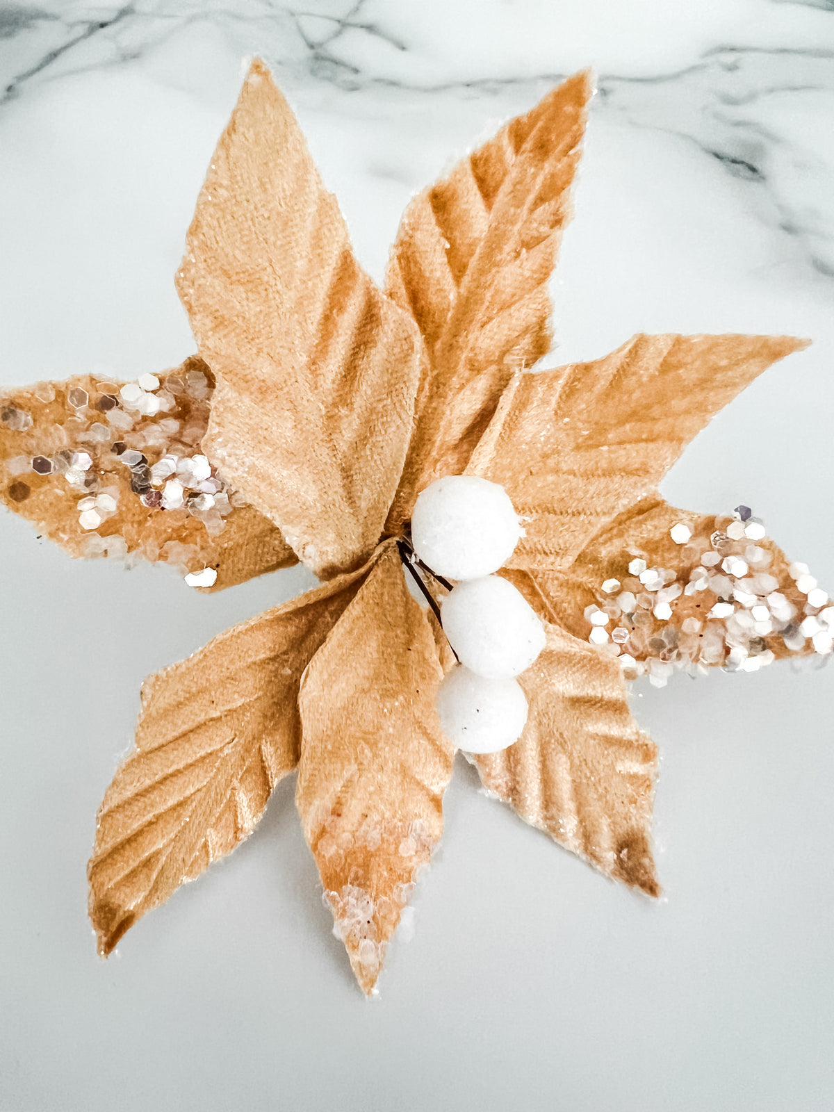 A decorative faux poinsettia flower ornament with metallic gold fabric leaves, white faux berries, and silver glitter accents, displayed on a light marble surface.