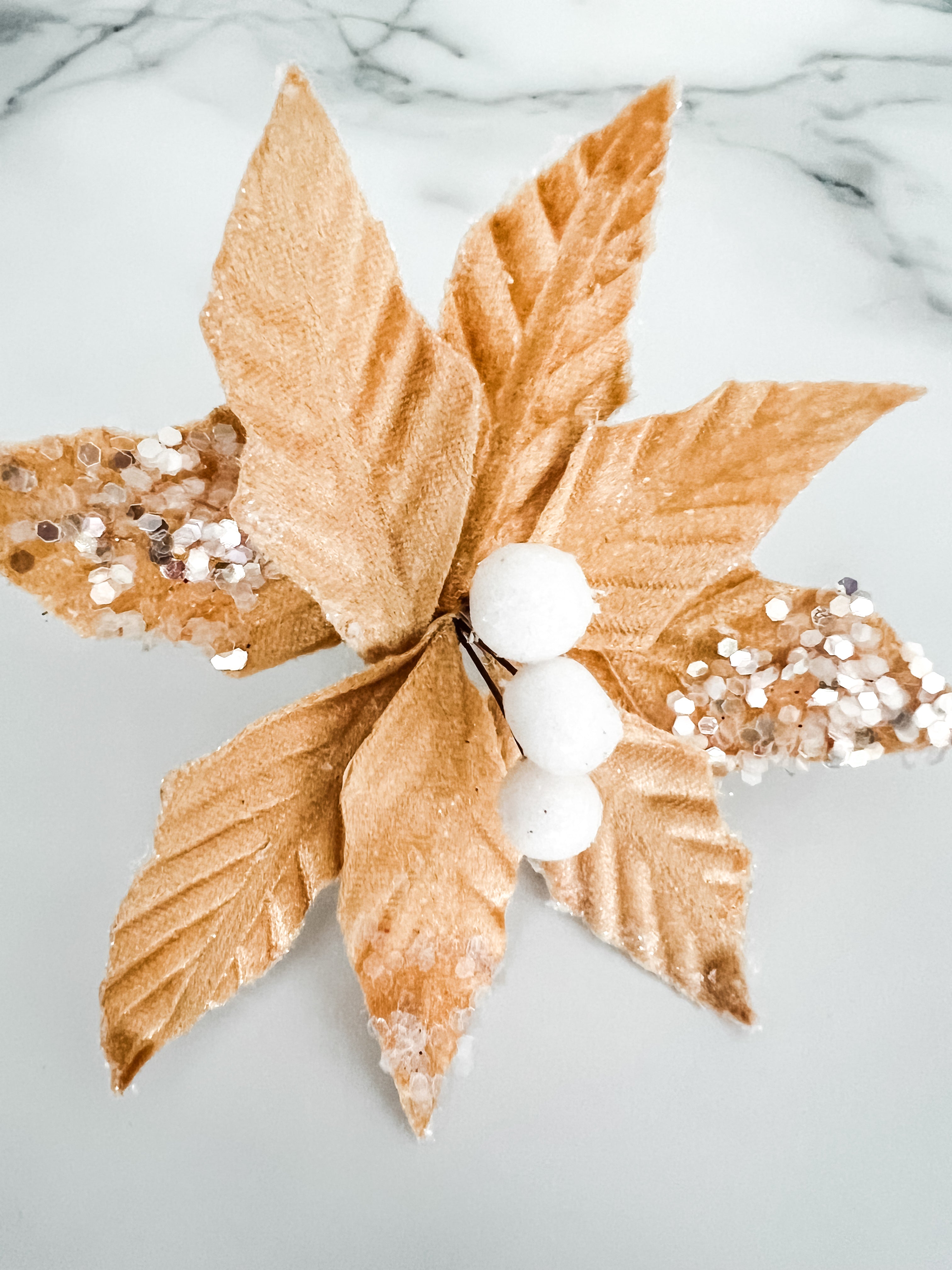 A decorative faux poinsettia flower ornament with metallic gold fabric leaves, white faux berries, and silver glitter accents, displayed on a light marble surface.