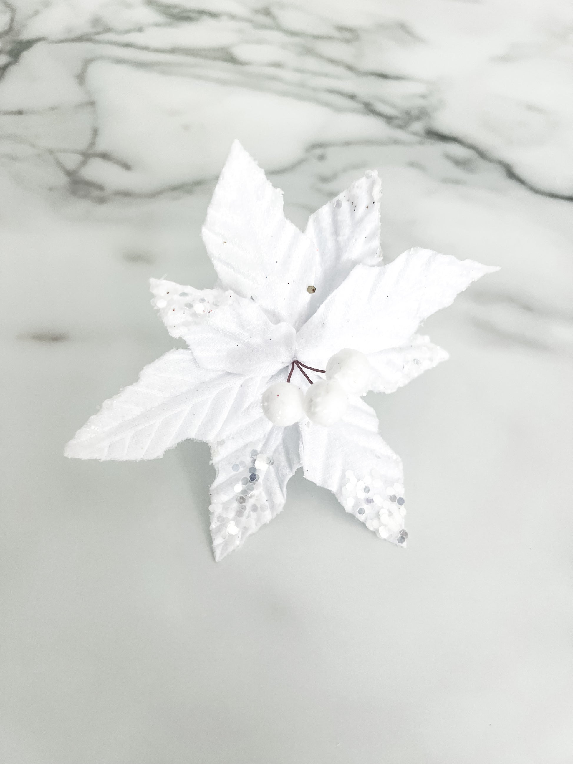 A white faux poinsettia decorative flower with textured leaves and small silver glitter accents, placed on a light marble surface.