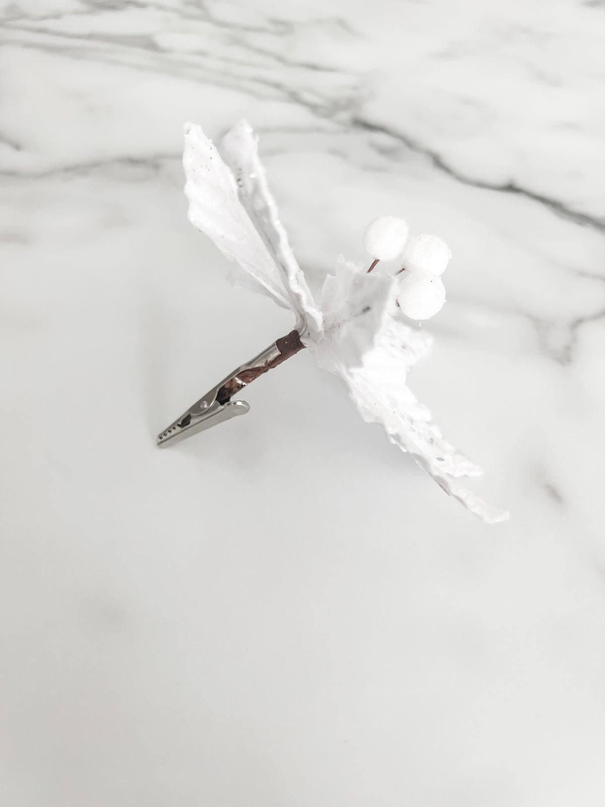 A white faux poinsettia decorative flower with textured leaves and small silver glitter accents and a silver clip, placed on a light marble surface.