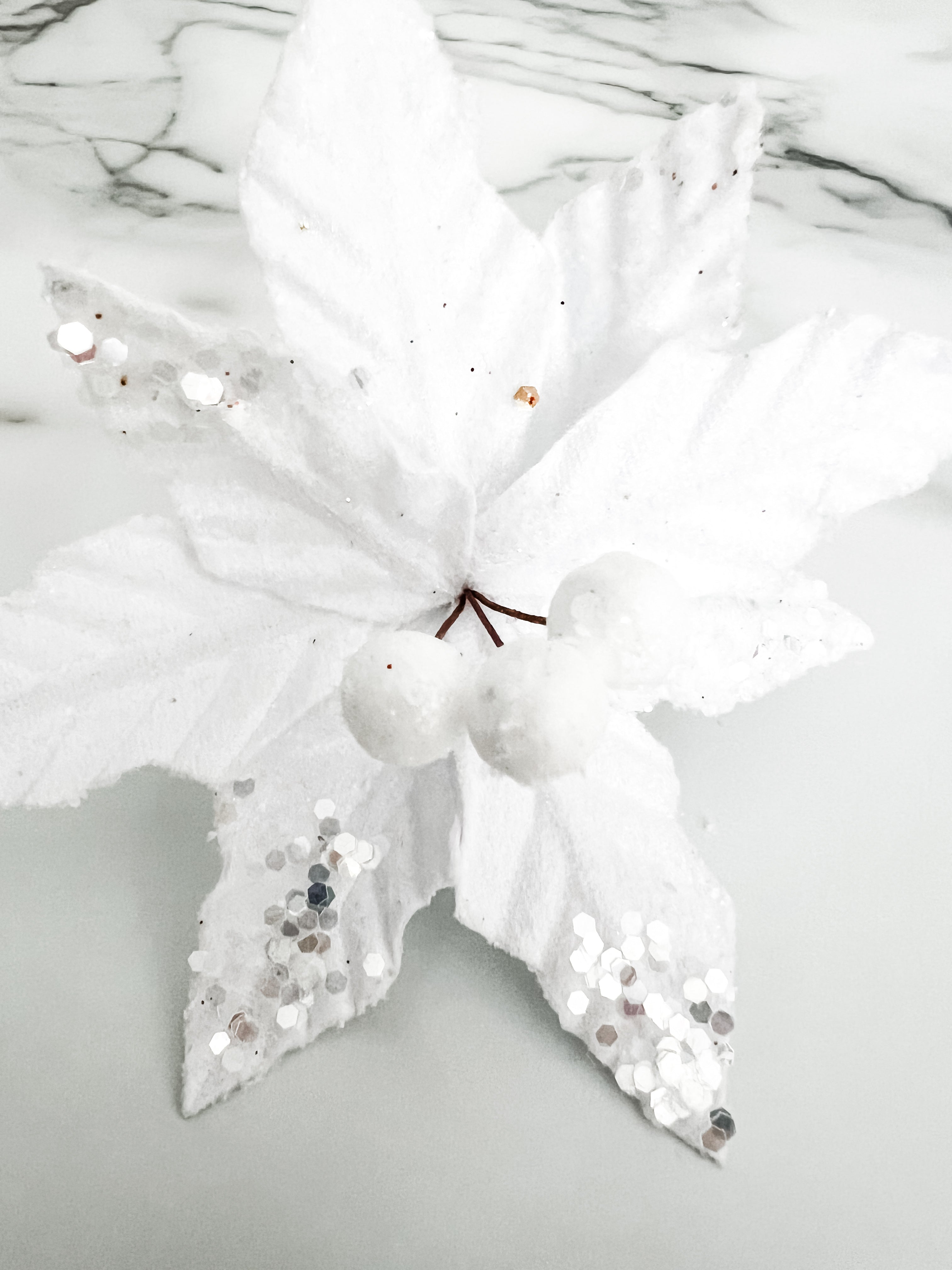 A white faux poinsettia decoration with textured fabric petals, silver sequins, and small white pom-pom berries on a light marble surface.
