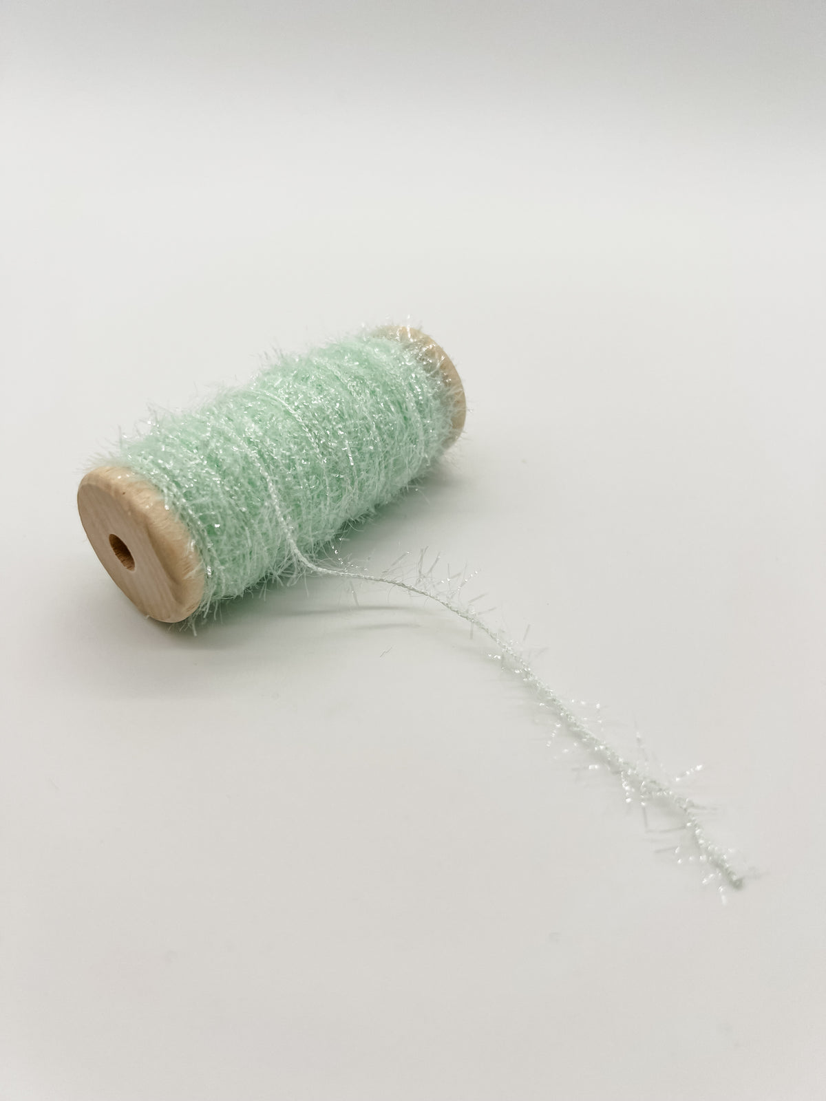 A small spool of fluffy, light mint green tinsel thread or decorative ribbon, shown on a plain light gray background.