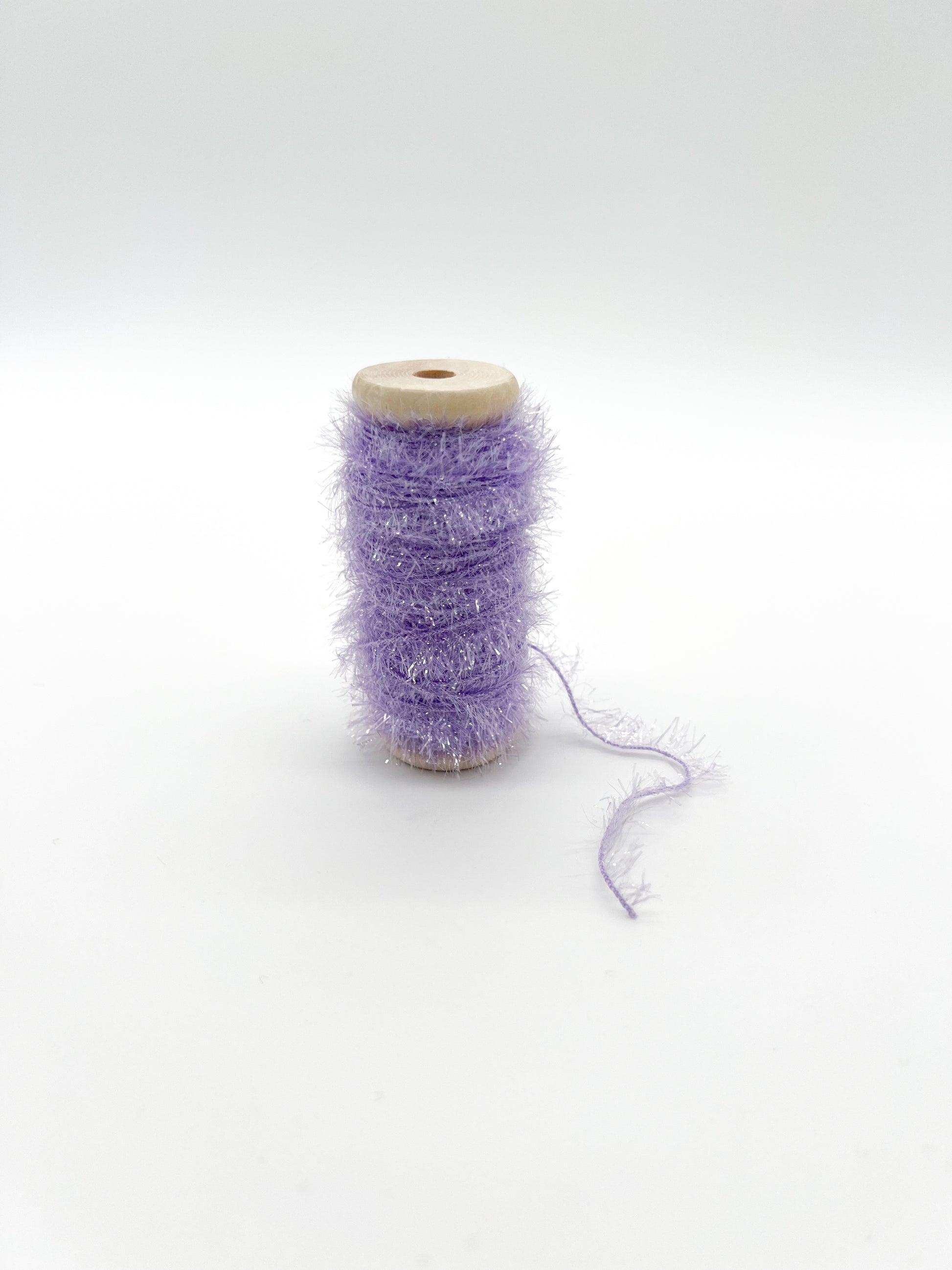 A spool of fluffy, shimmery lavender purple thread or ribbon on a light wood core, set against a seamless white studio background.