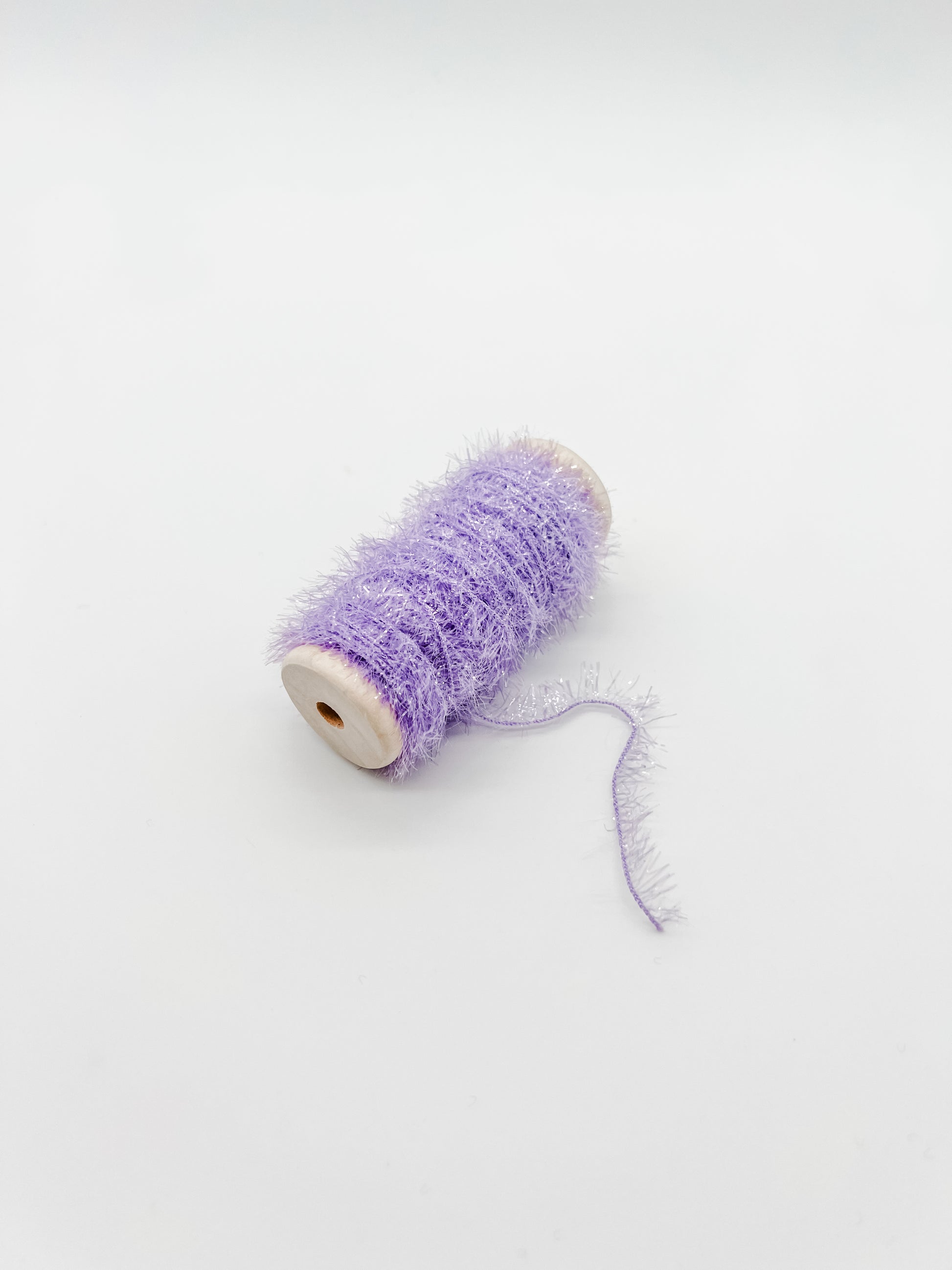 A spool of fluffy, shimmery lavender purple tinsel thread or craft yarn, shown on a plain white background.