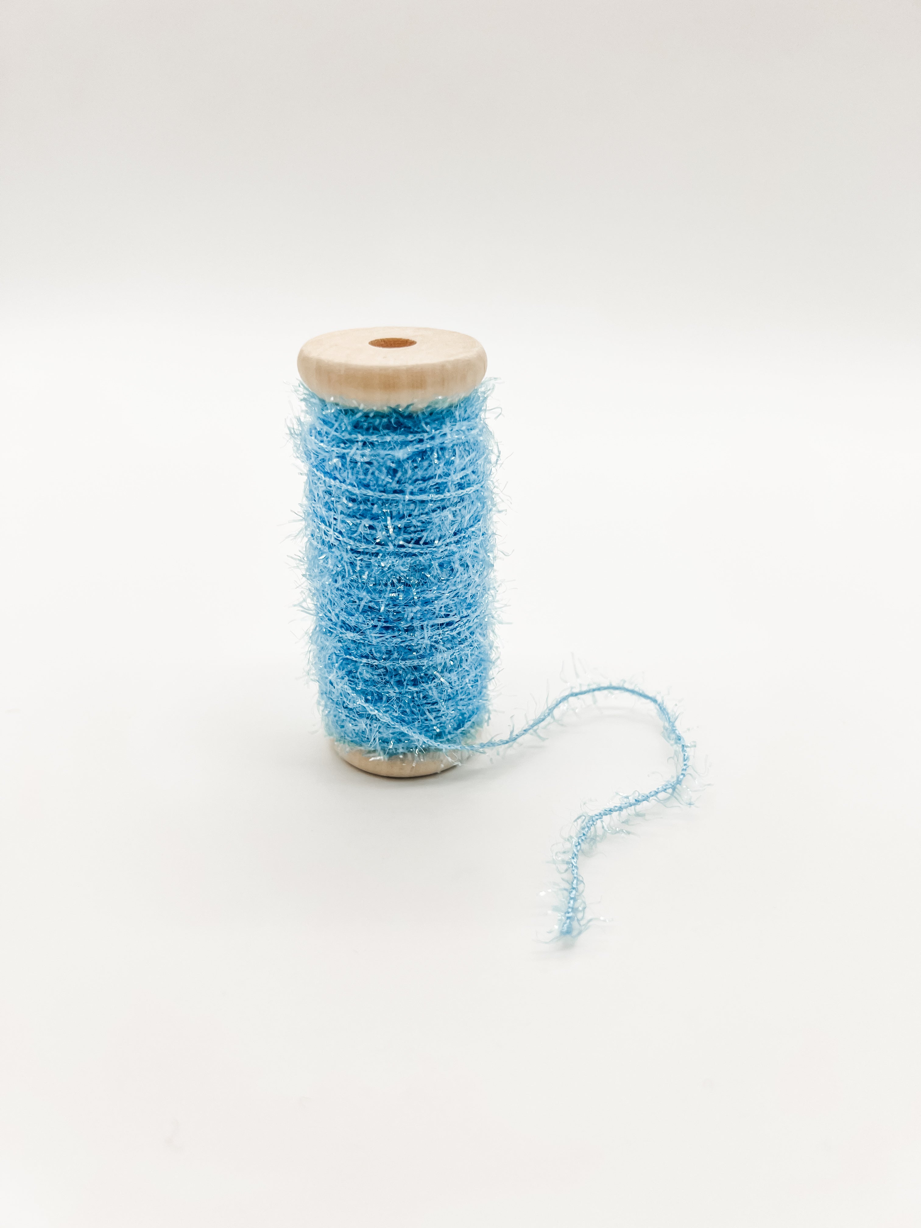 A spool of light blue fuzzy tinsel craft thread with a natural wood core, shown on a seamless white background.