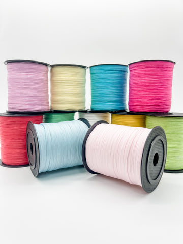 Eleven spools of paper raffia arranged in two rows on a clean white background. The front row includes light blue, pale pink, and lime green spools, while the back row shows lavender, cream, teal, and hot pink spools and other colors.