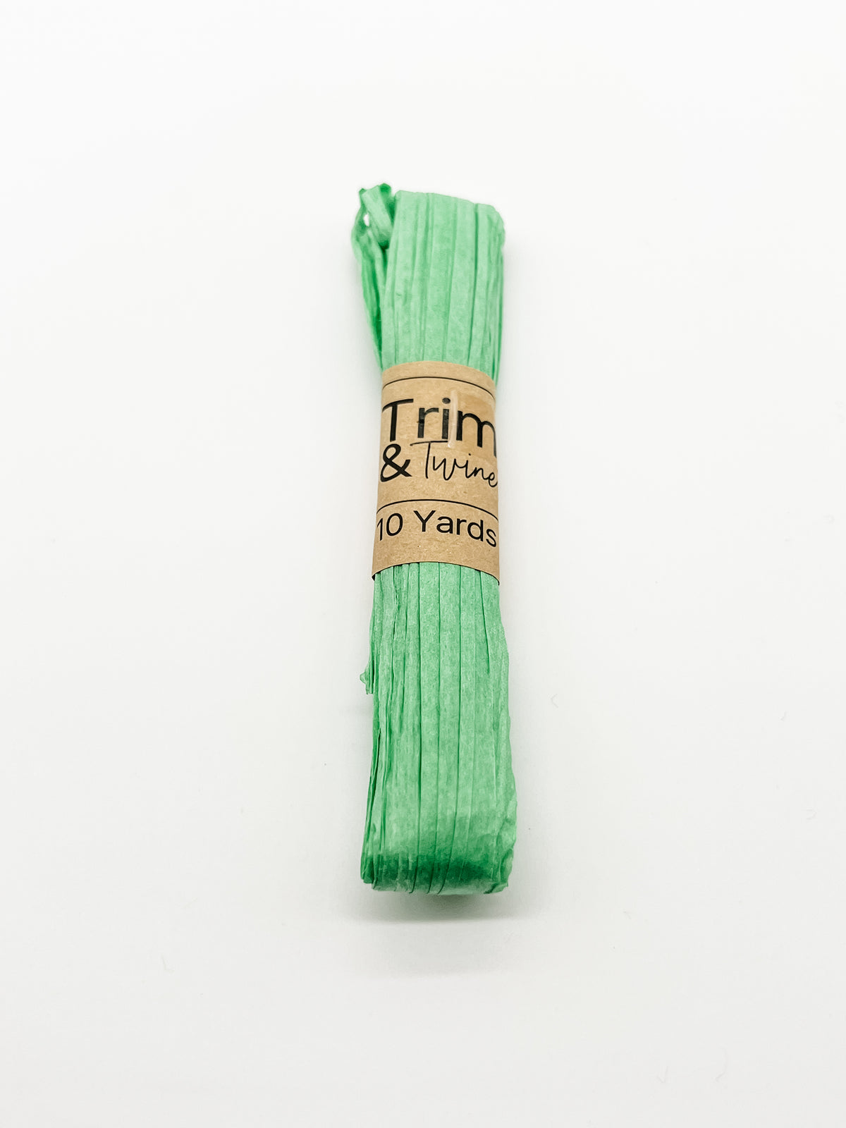 A roll of green raffia wrapped with a brown paper band that reads 'Trim & Twine' and '10 Yards', photographed on a plain white background.