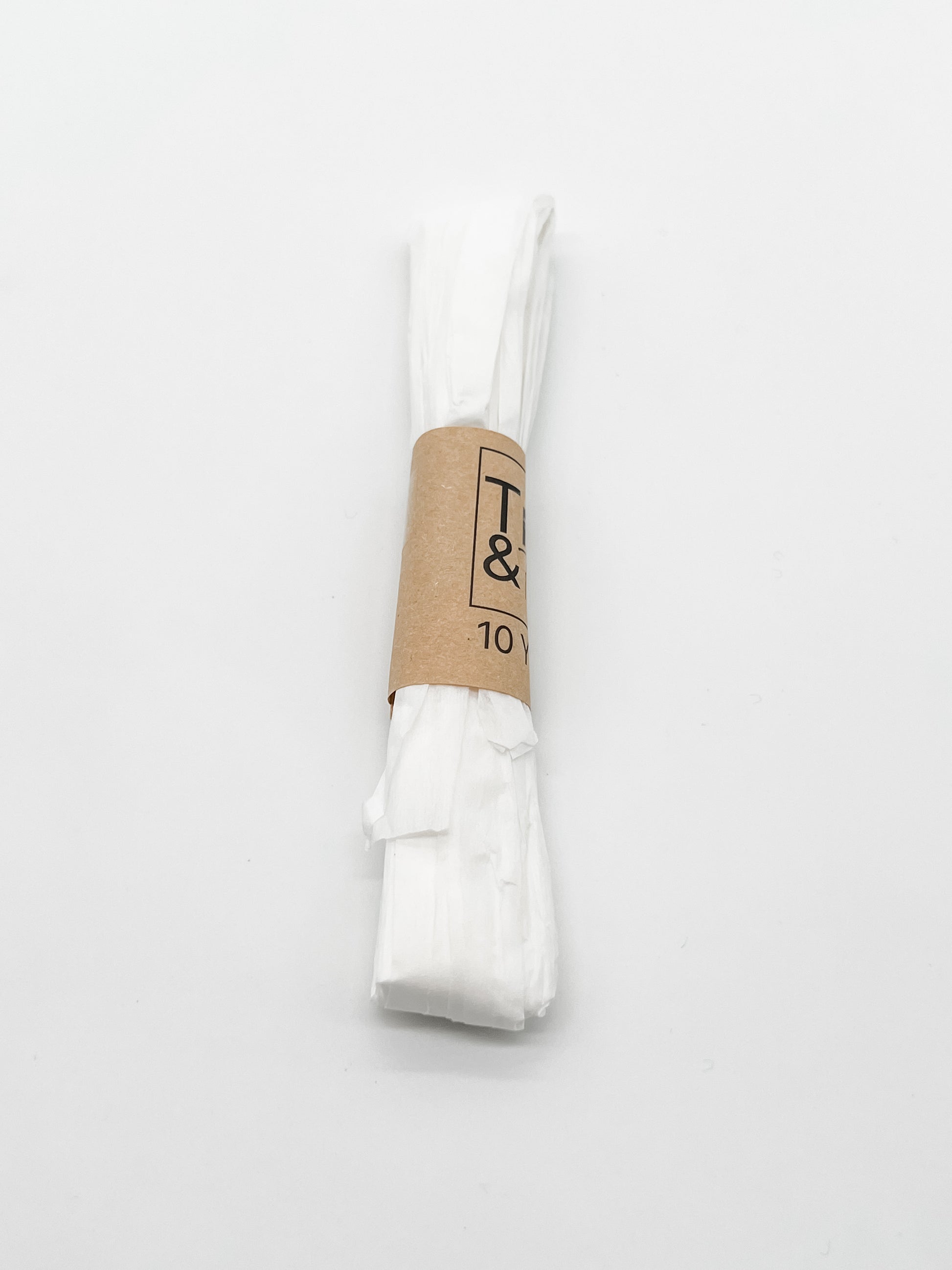 A roll of white raffia wrapped with a brown paper band that reads 'Trim & Twine' and '10 Yards', photographed on a plain white background.