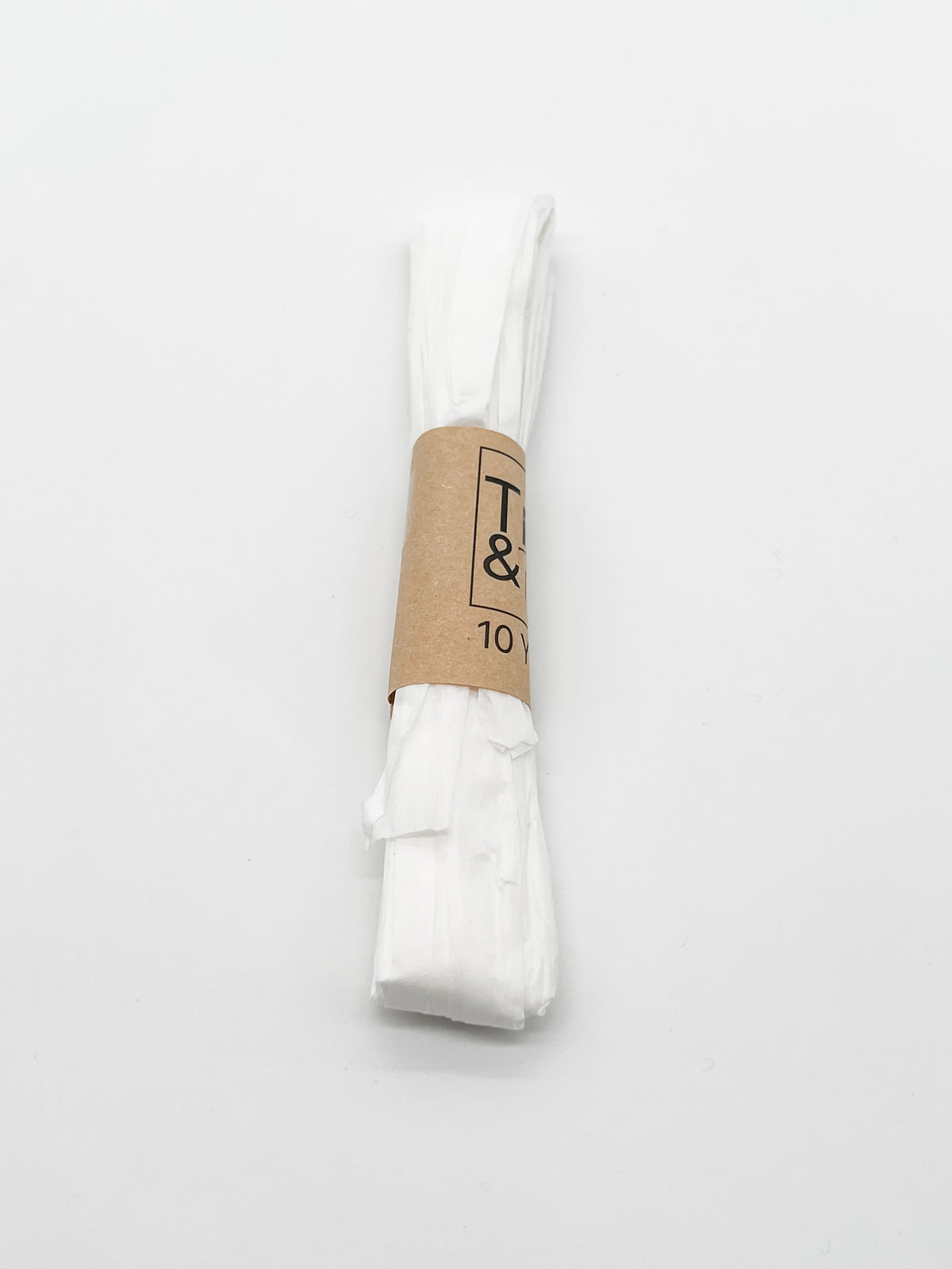 A roll of white raffia wrapped with a brown paper band that reads 'Trim & Twine' and '10 Yards', photographed on a plain white background.