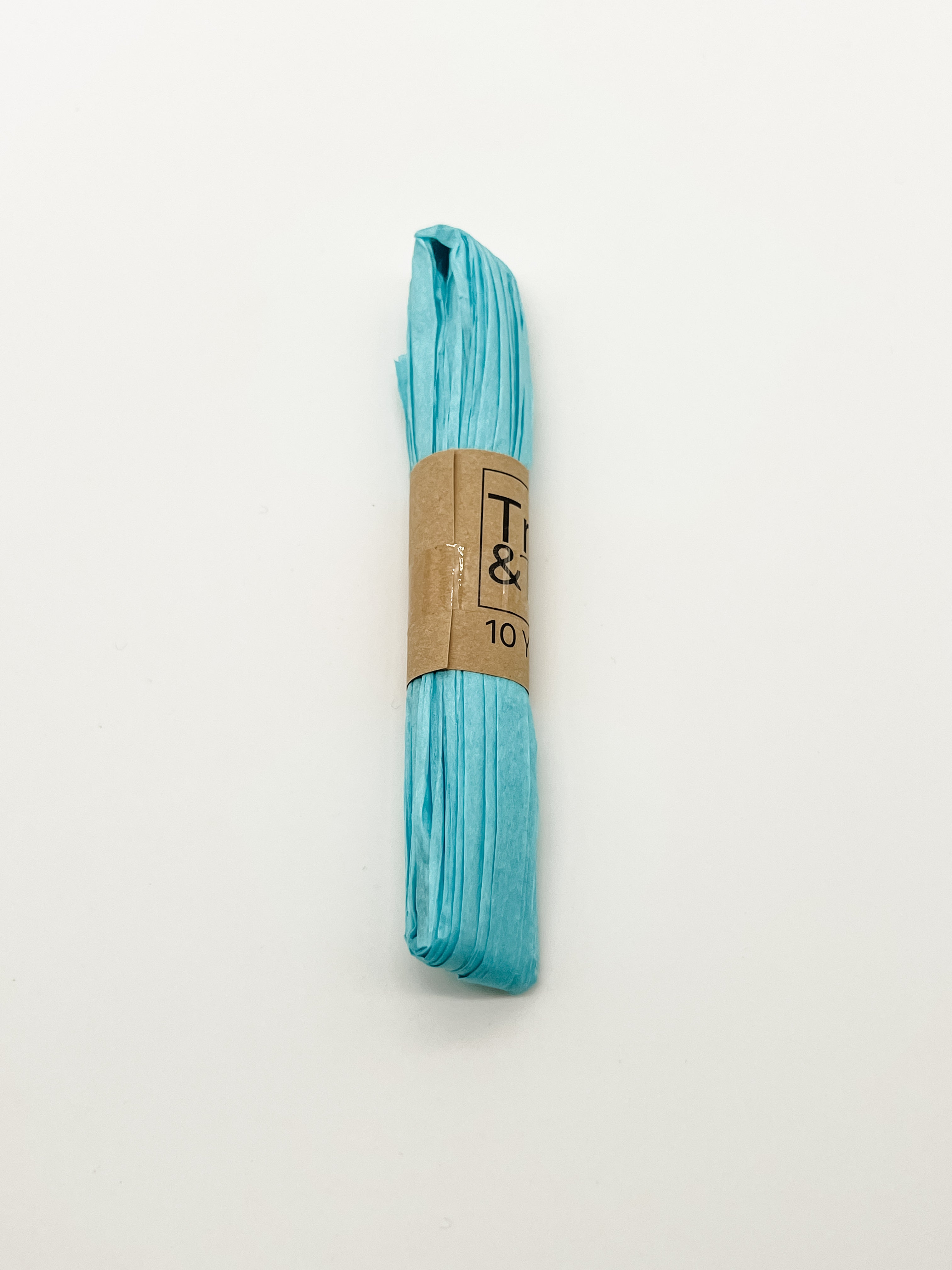 A roll of blue raffia wrapped with a brown paper band that reads 'Trim & Twine' and '10 Yards', photographed on a plain white background.