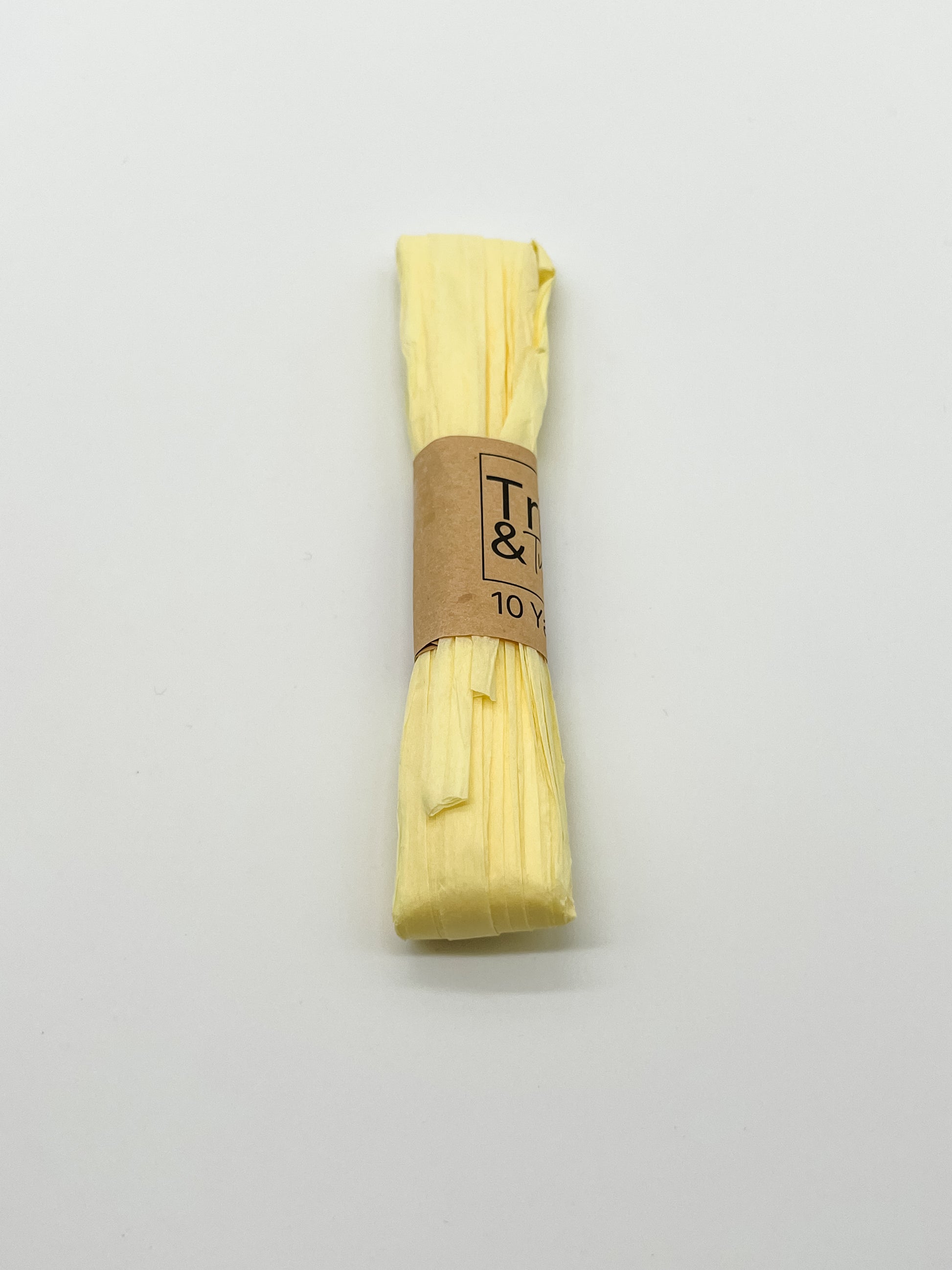A roll of pale yellow raffia wrapped with a brown paper band that reads 'Trim & Twine' and '10 Yards', photographed on a plain white background.