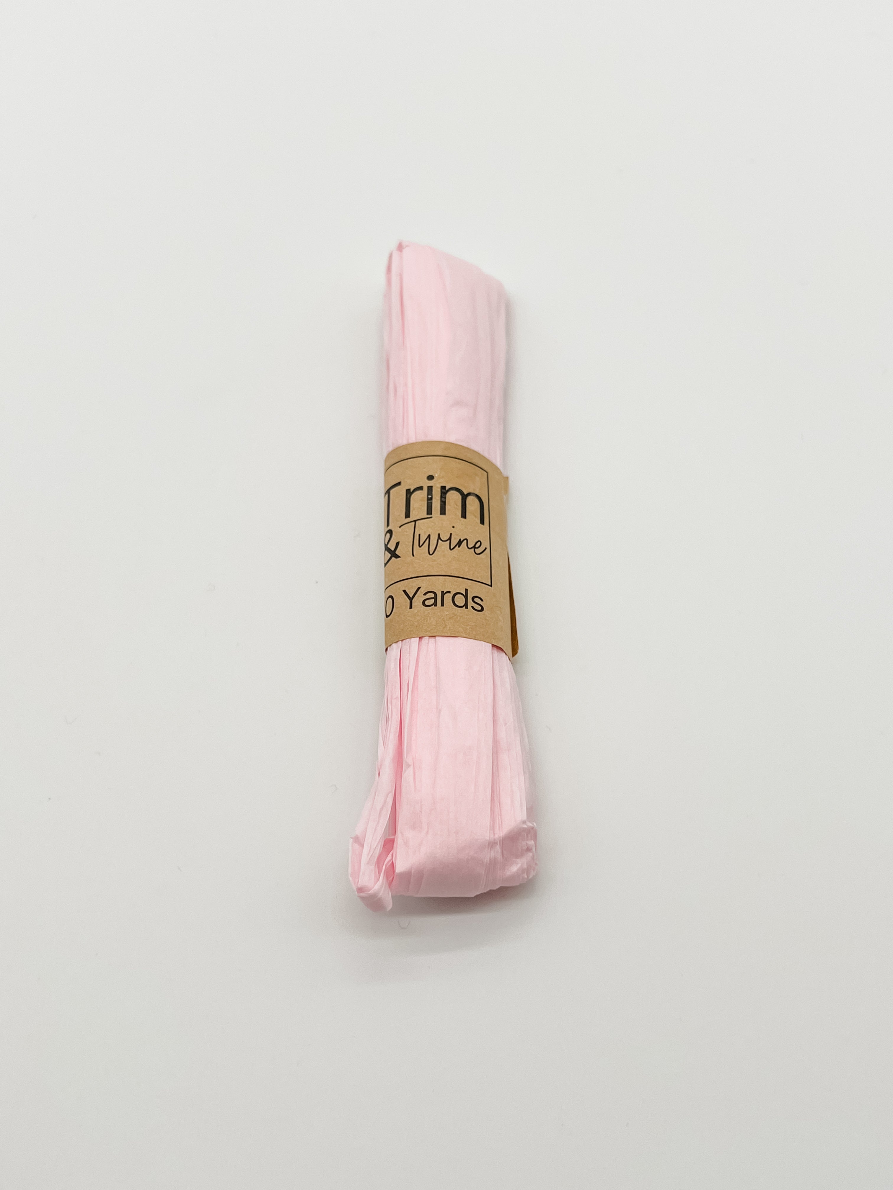 A roll of pale pink raffia wrapped with a brown paper band that reads 'Trim & Twine' and '10 Yards', photographed on a plain white background.
