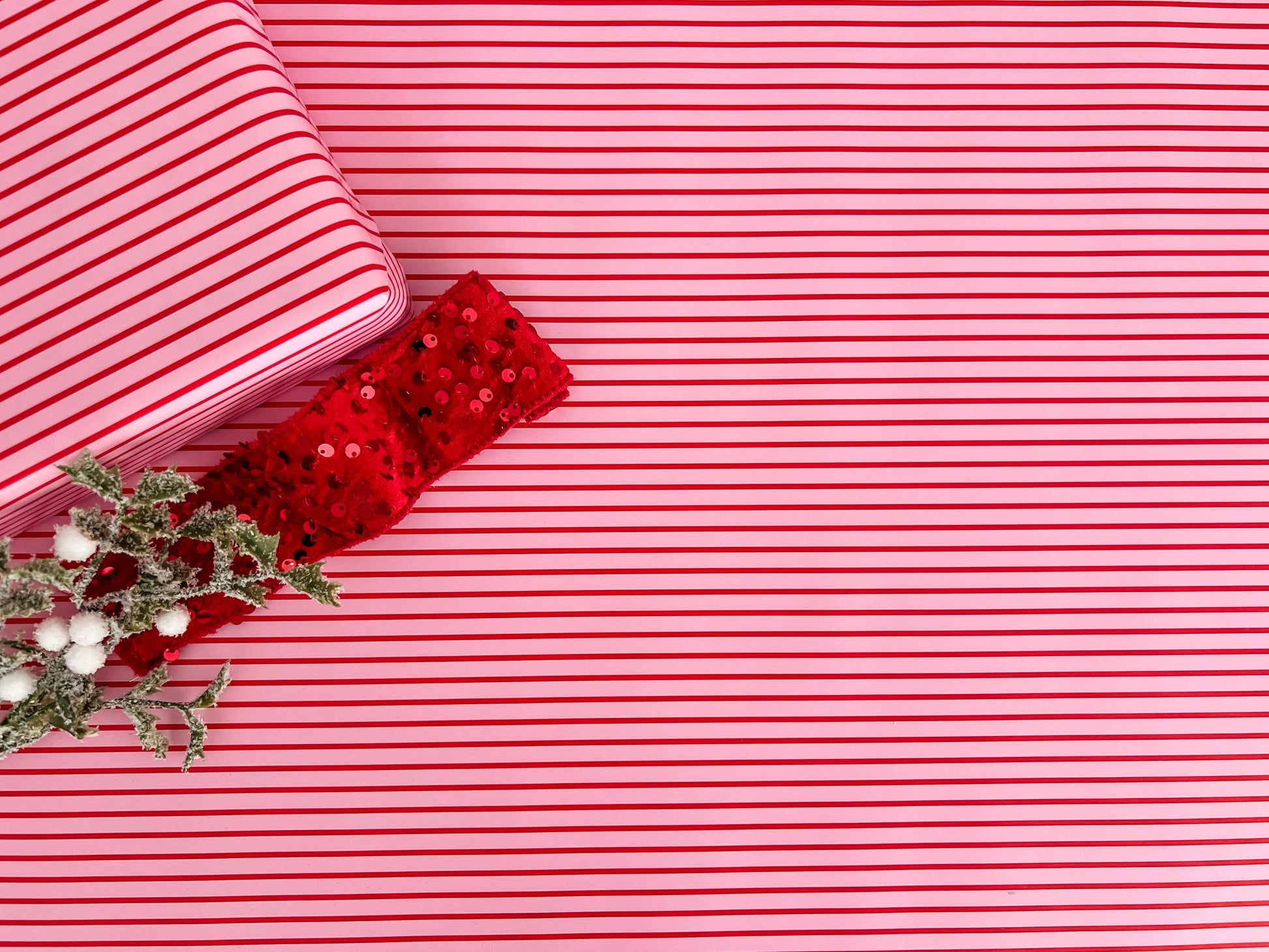 Red and pink striped wrapping paper with a red sequin ribbon and greenery on a matching background