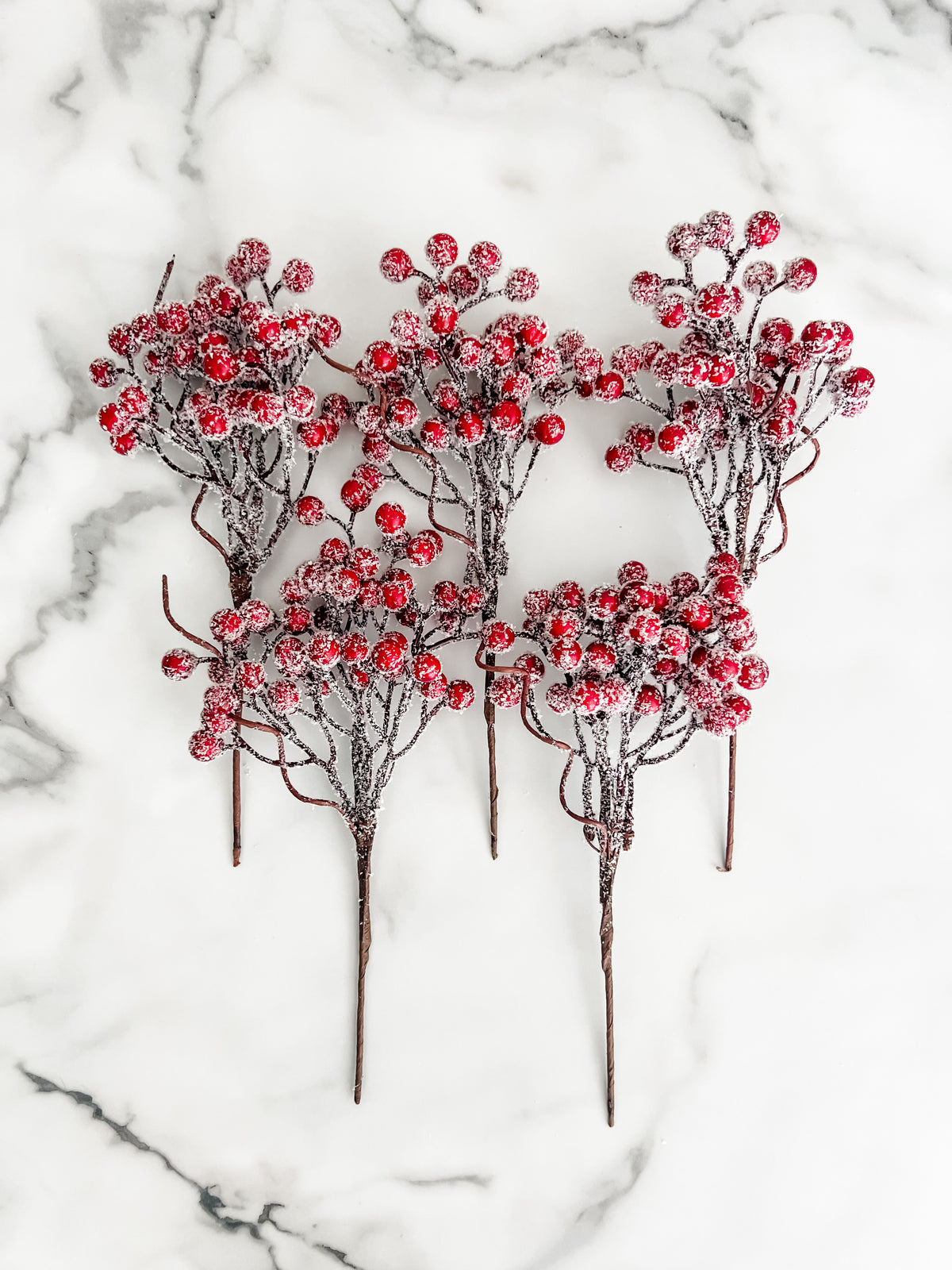 Five artificial frosted berry stems with red berries and dark brown branches, arranged on a white marble surface.
