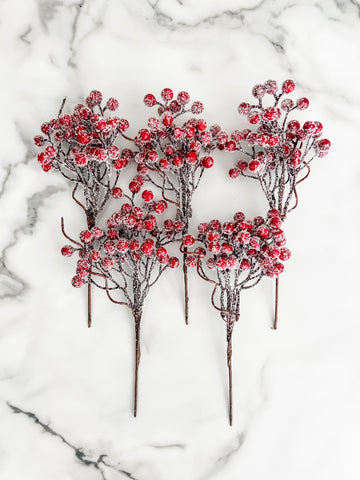 Five artificial frosted berry stems with red berries and dark brown branches, arranged on a white marble surface.