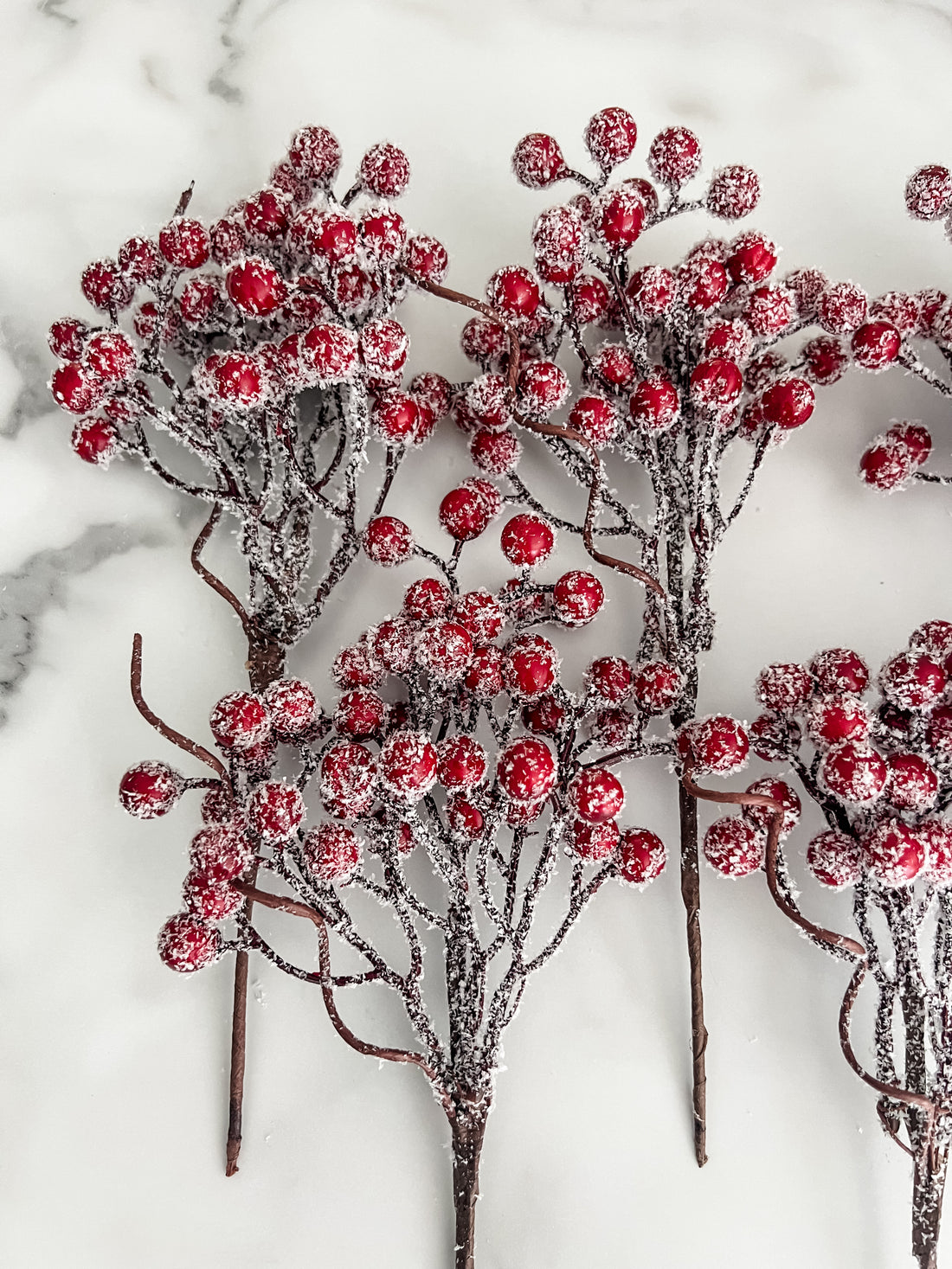 Five artificial frosted berry stems with red berries and dark brown branches, arranged on a white marble surface.