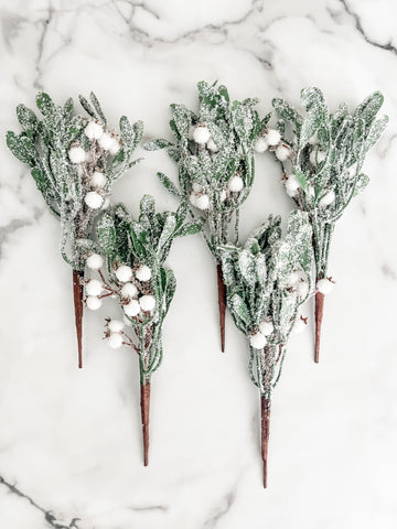 Five artificial frosted greenery stems with white berries and brown wooden picks, arranged on a white marble surface.