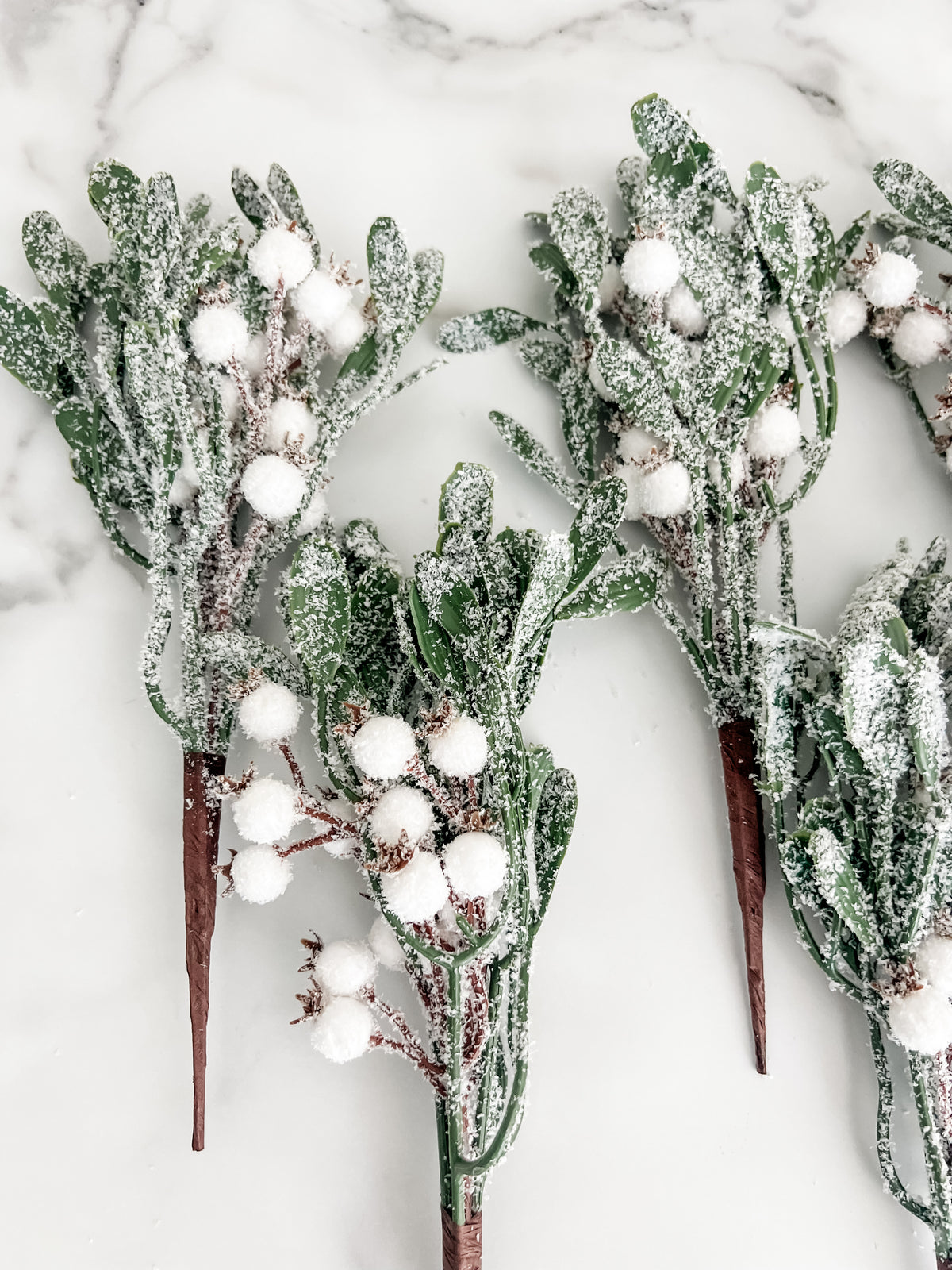 Frosted green foliage stems with white pom-pom berries, arranged on a white marble surface.