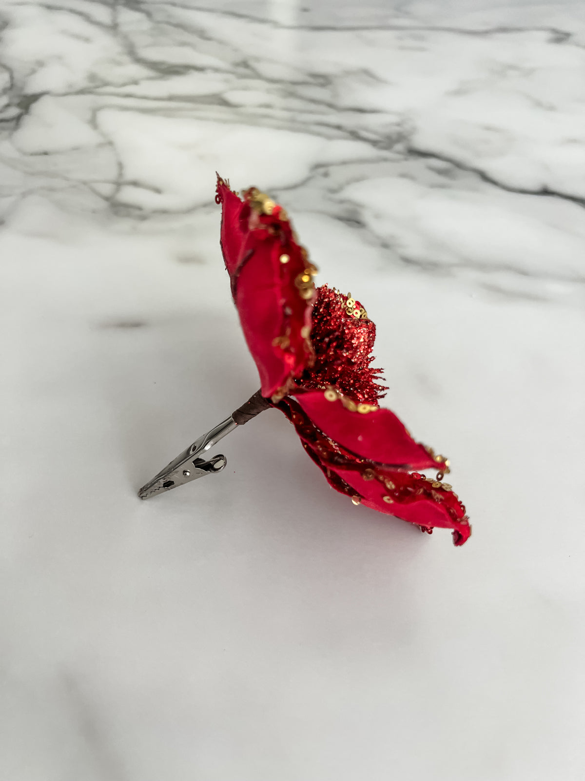A red fabric flower clip with glittery red center and gold sequin accents, photographed on a white marble surface.